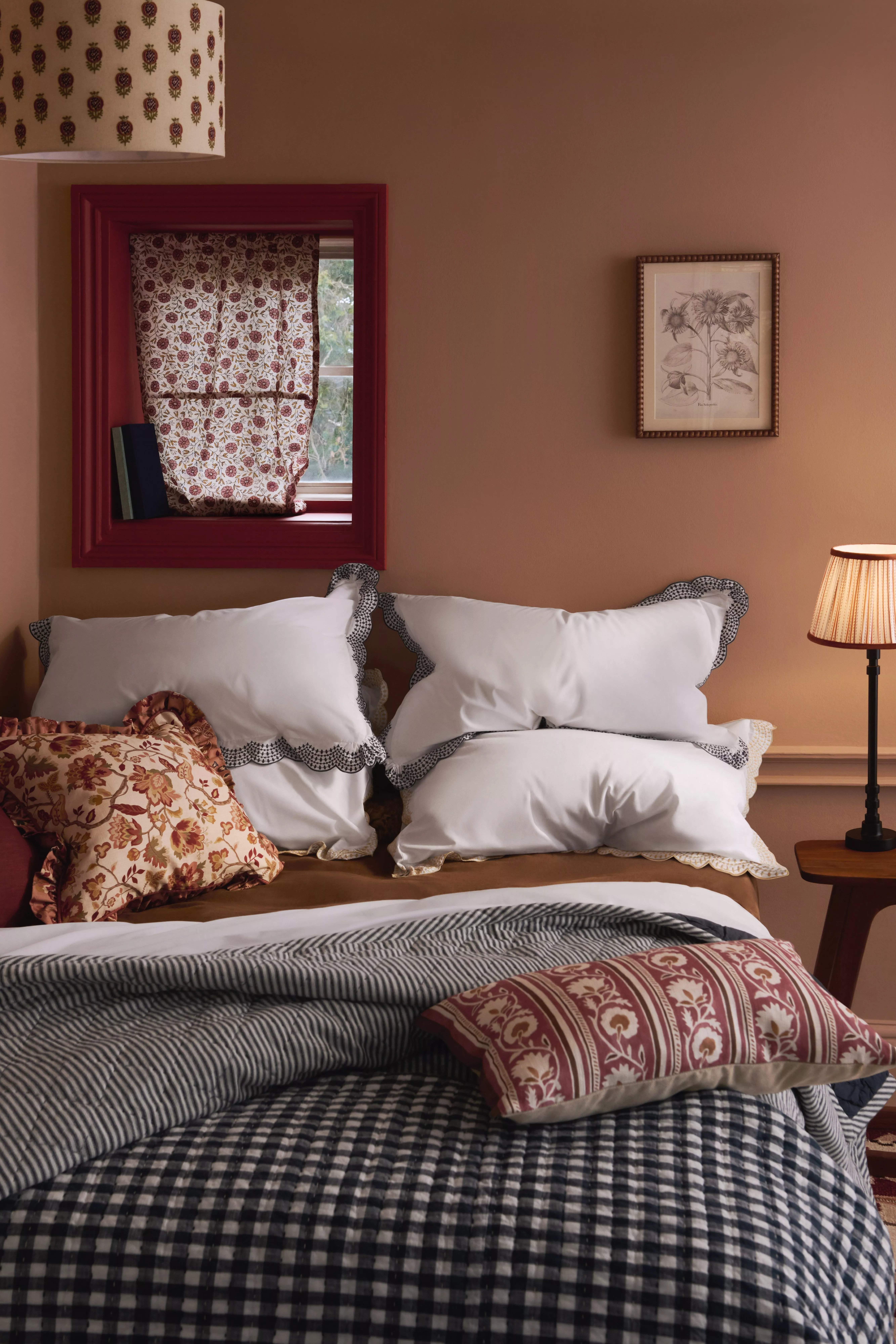 Cosy bedroom with patterned cushions, gingham duvet, red framed window, floral curtain and warm bedside lamp.