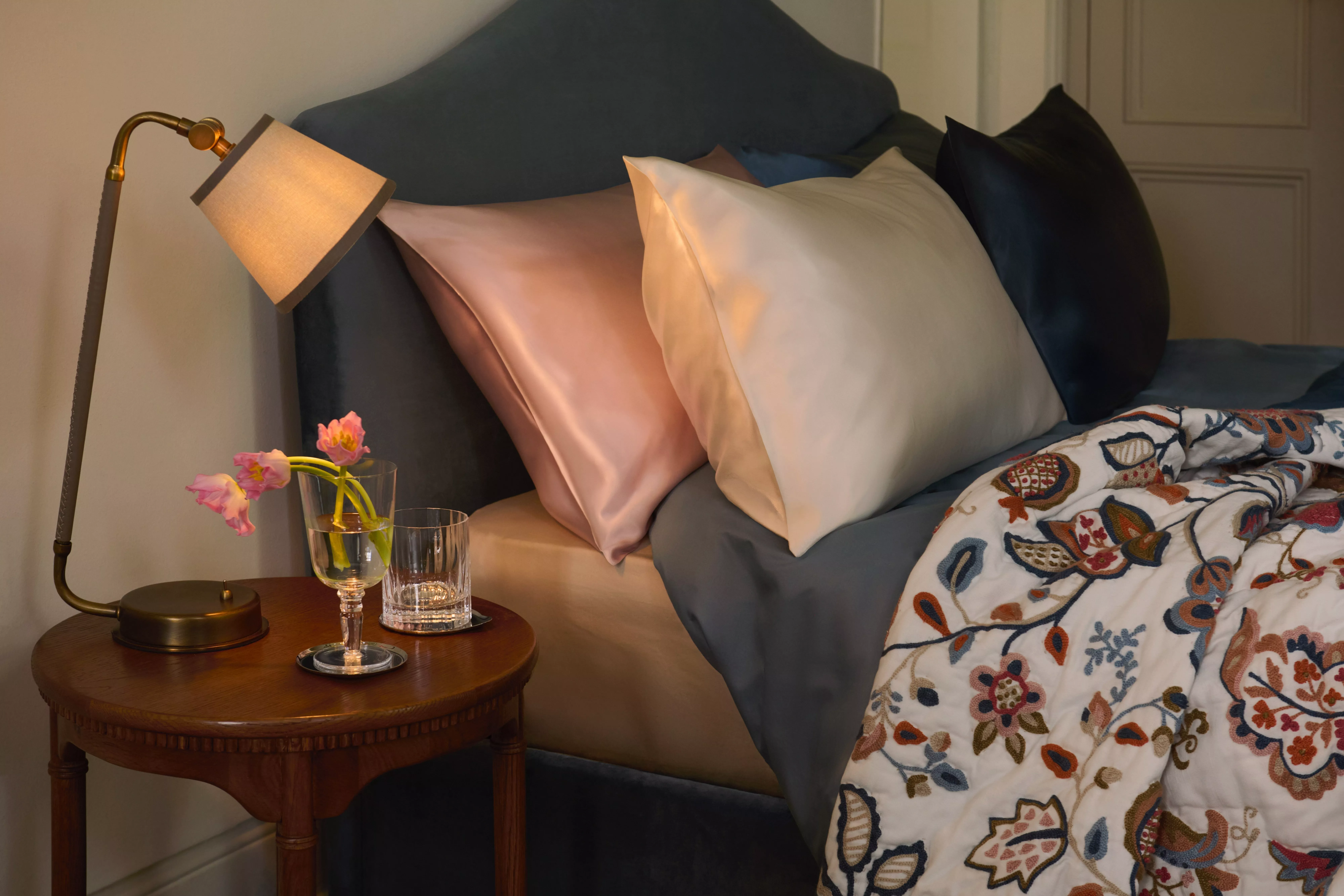 Cosy bedroom with velvet headboard, satin pillows, embroidered duvet, and bedside lamp with flowers on table