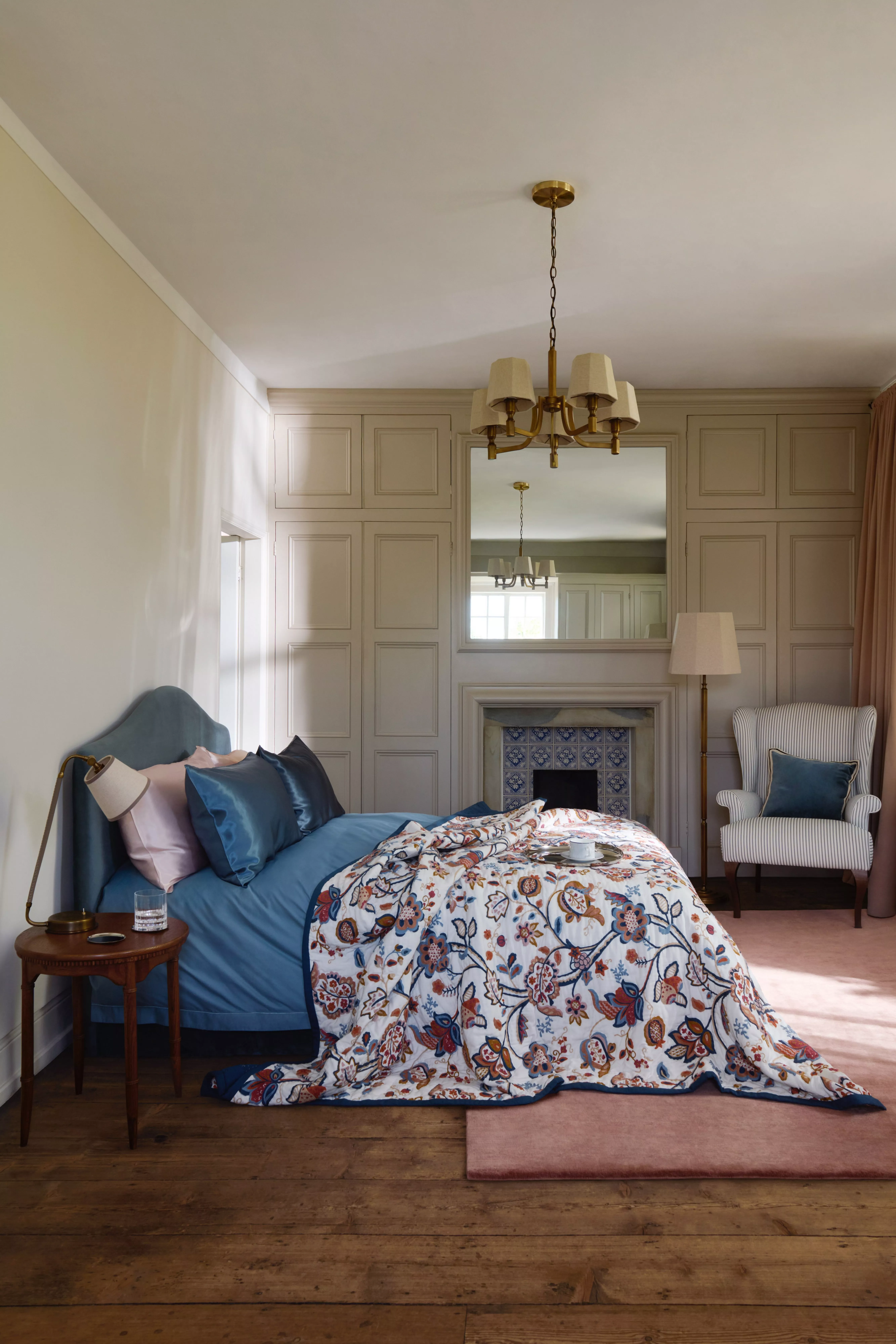 Cosy bedroom interior with blue upholstered bed, floral duvet, panelled walls, fireplace and brass chandelier lighting