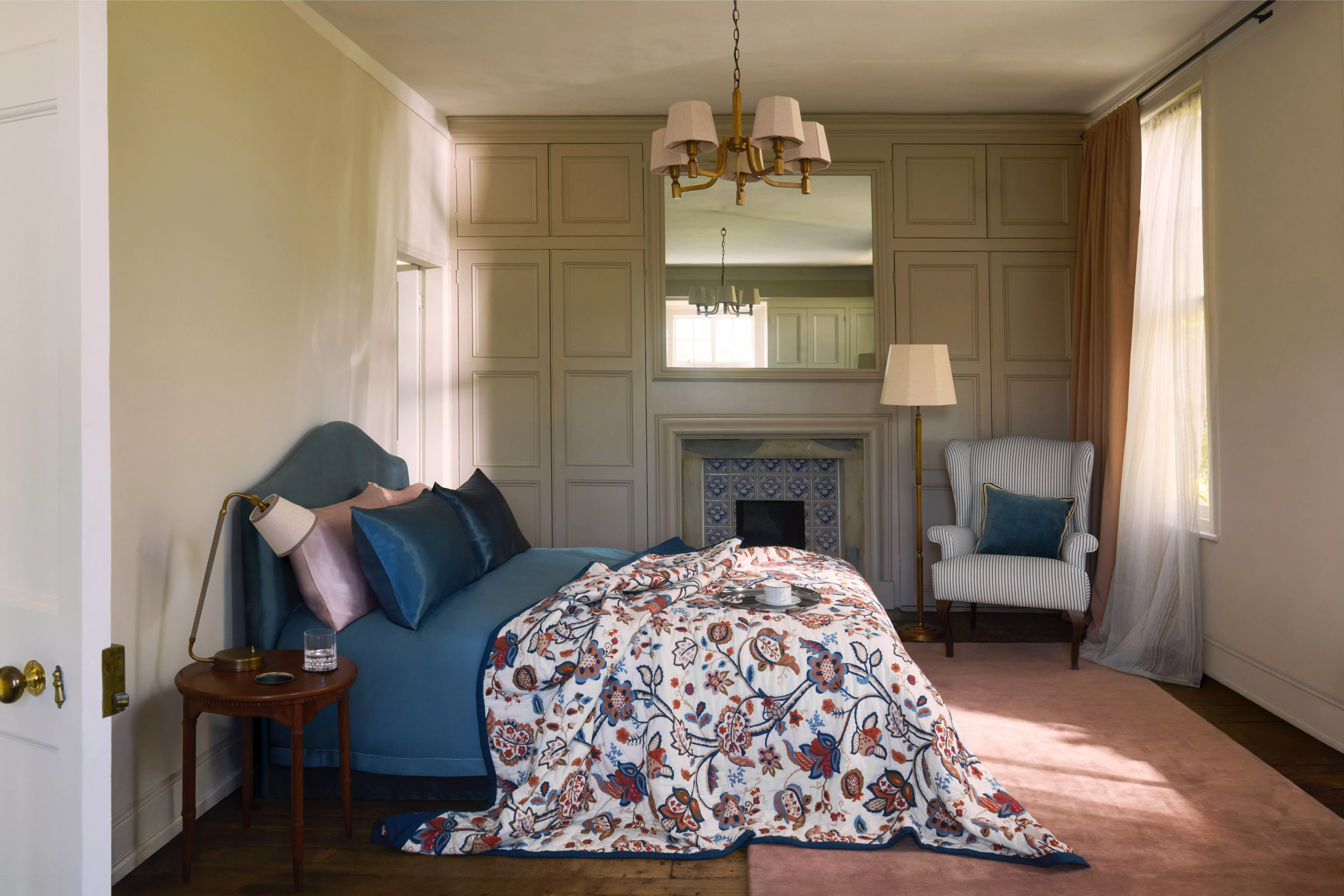 Elegant bedroom interior with blue upholstered bed, floral duvet, panelled walls, fireplace mirror and armchair by window