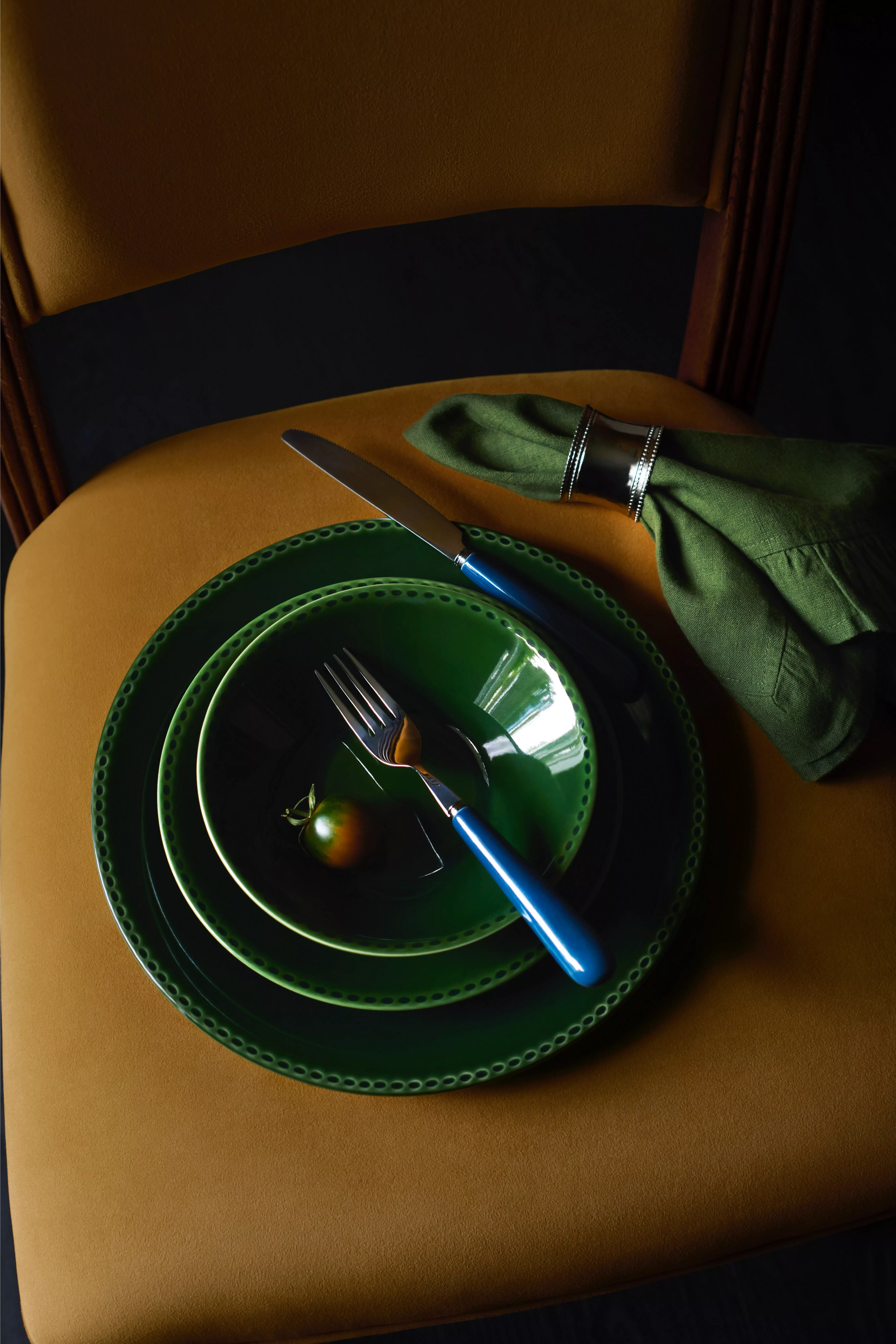 Green dinner plates with blue-handled cutlery and linen napkin on mustard chair, stylish table setting close-up