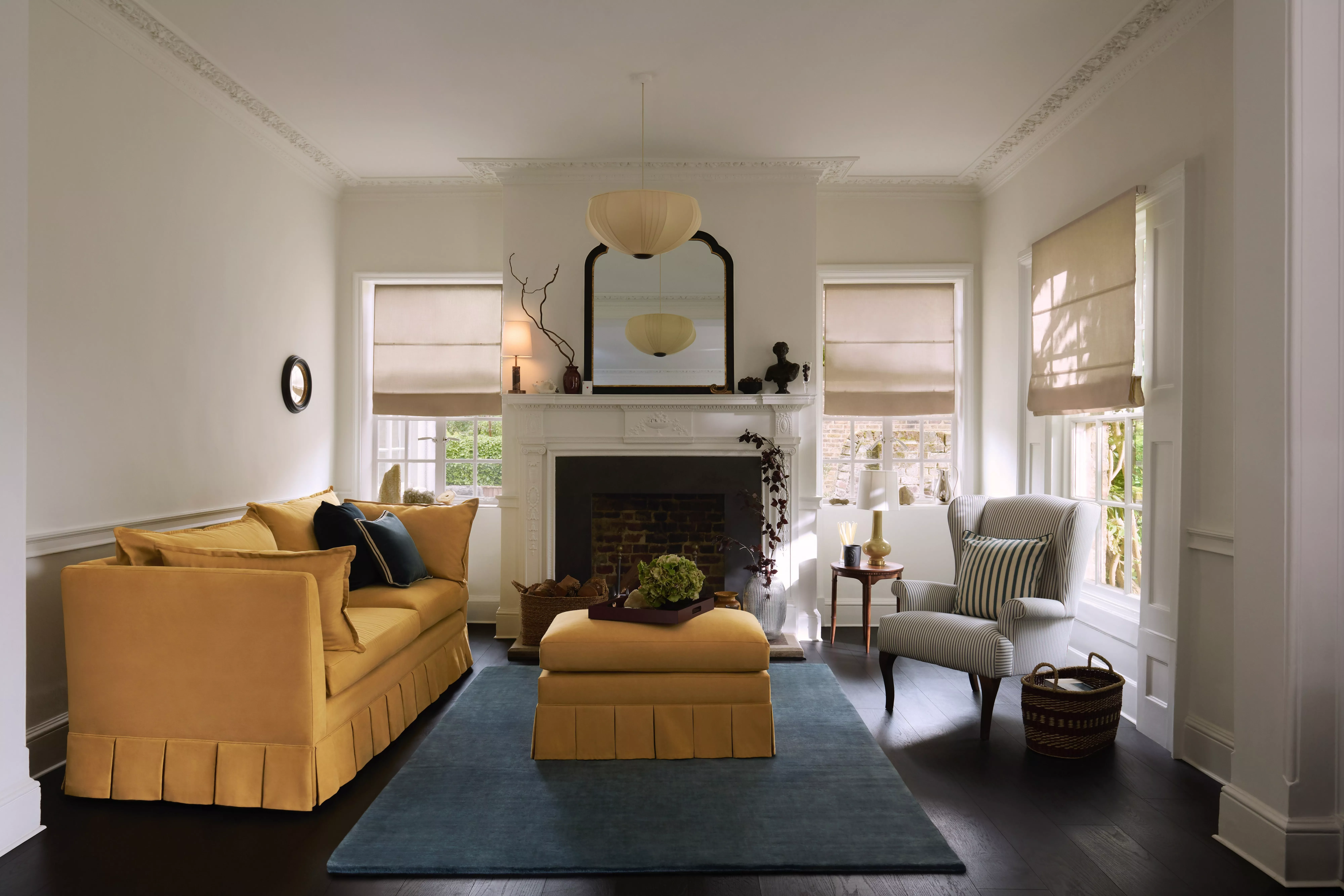 Bright traditional living room with yellow sofa, blue rug, white fireplace and Roman blinds by windows