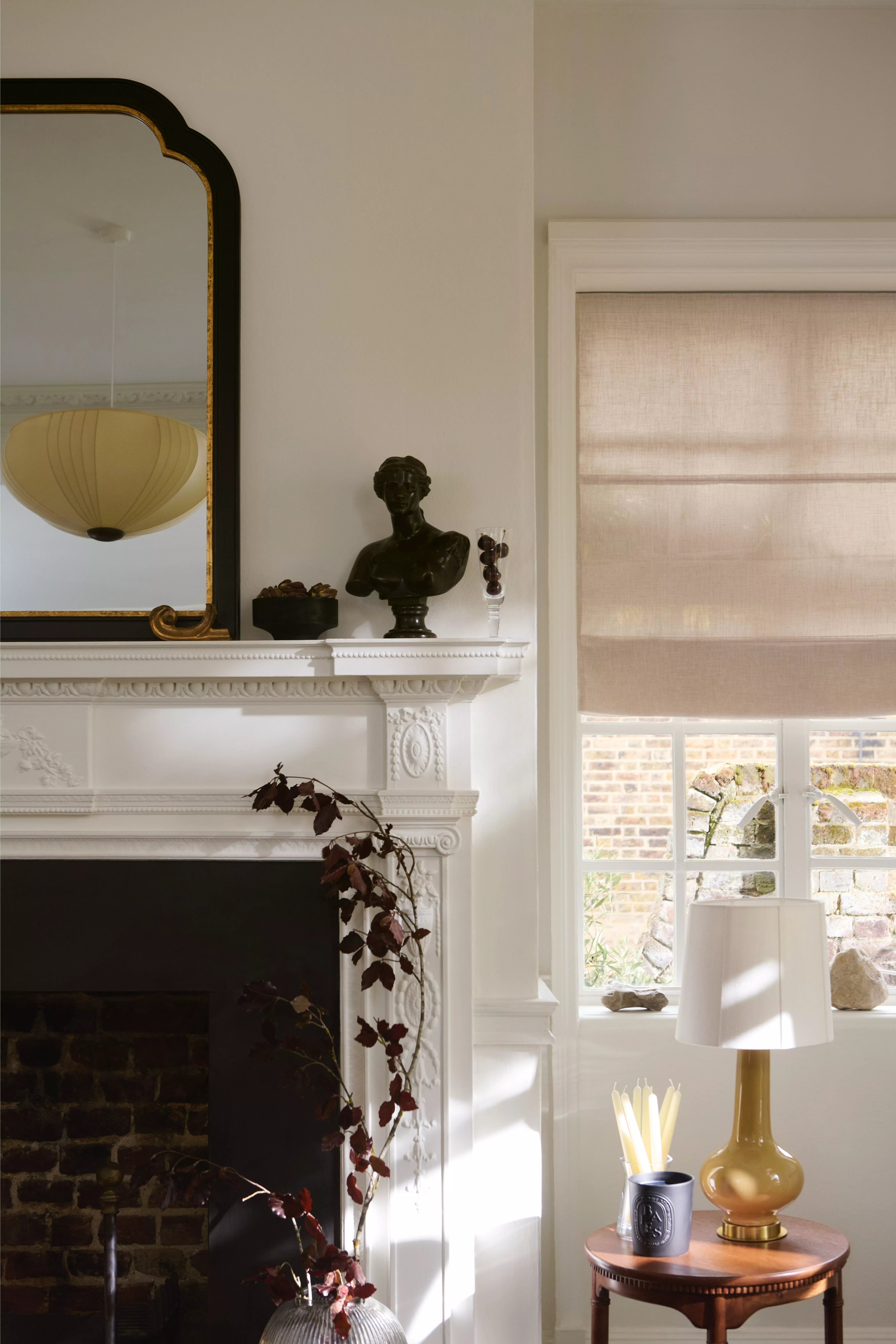 Bright living room with ornate fireplace, vintage mirror, Roman blind window, and amber table lamp on side table
