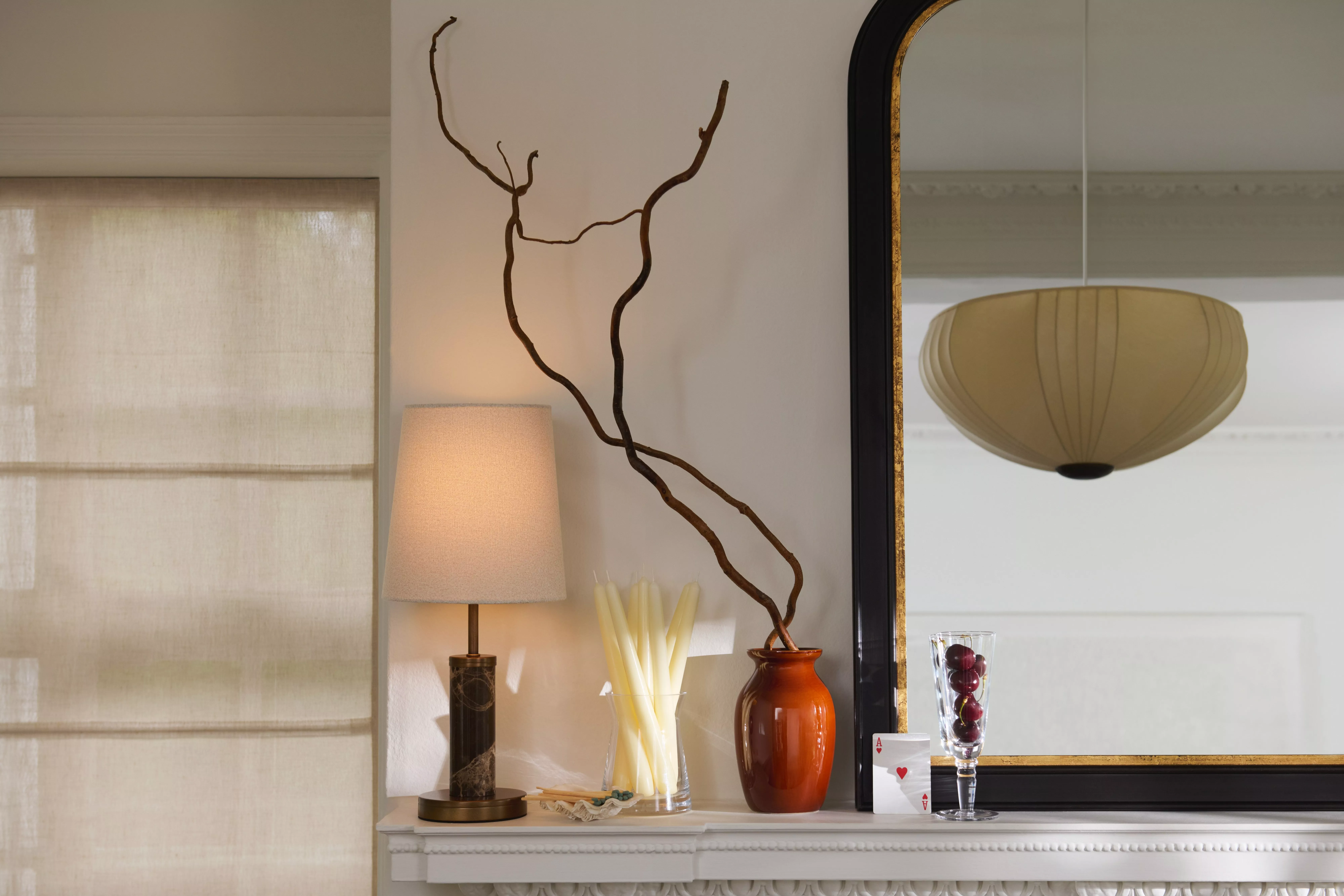 Elegant living room mantel with table lamp, tall vase branches, candles, large mirror and glass of grapes