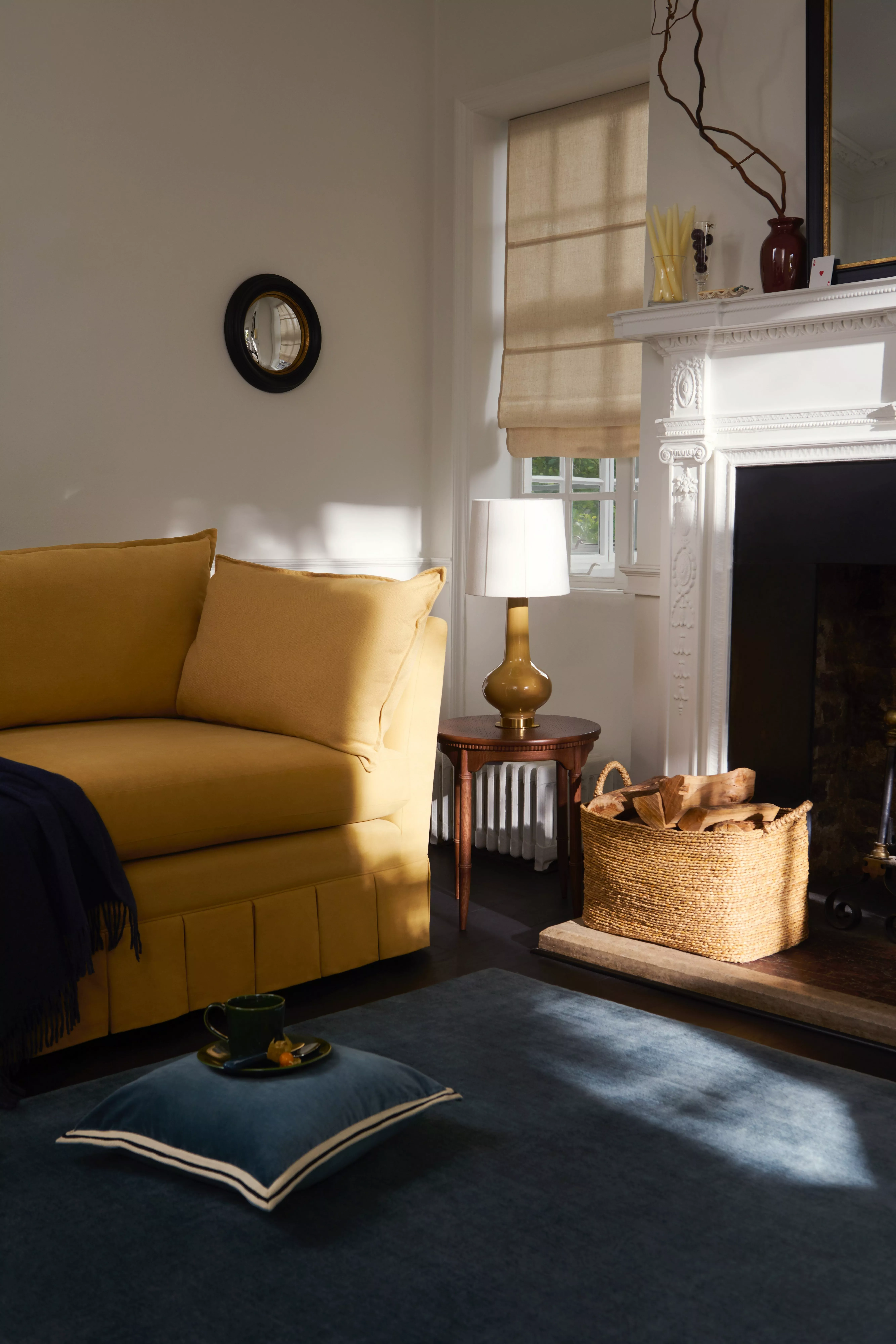 Cosy living room with mustard sofa, white fireplace, woven log basket, table lamp and beige Roman blind.
