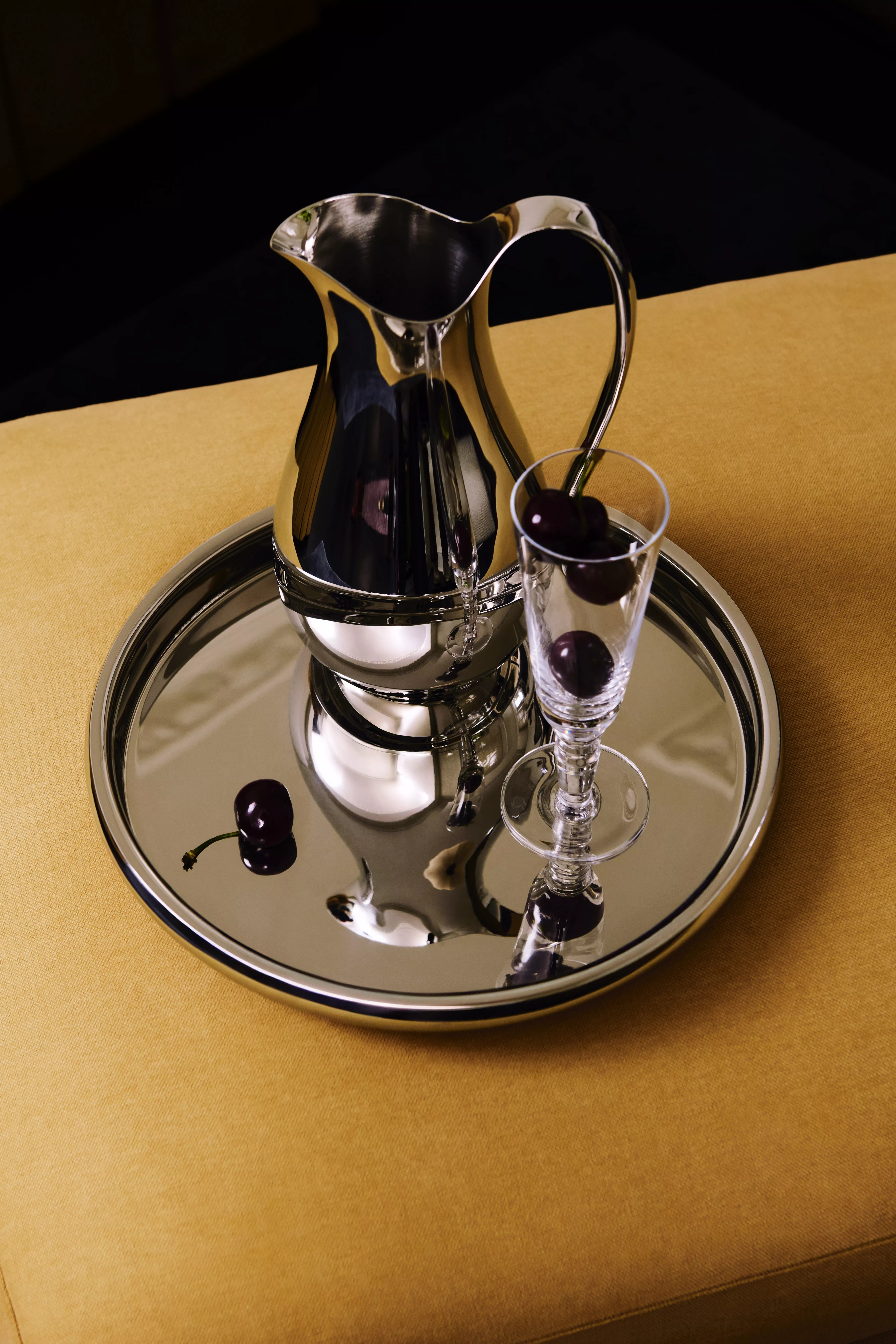 Polished silver pitcher and mirrored serving tray with glass of black cherries on mustard tablecloth