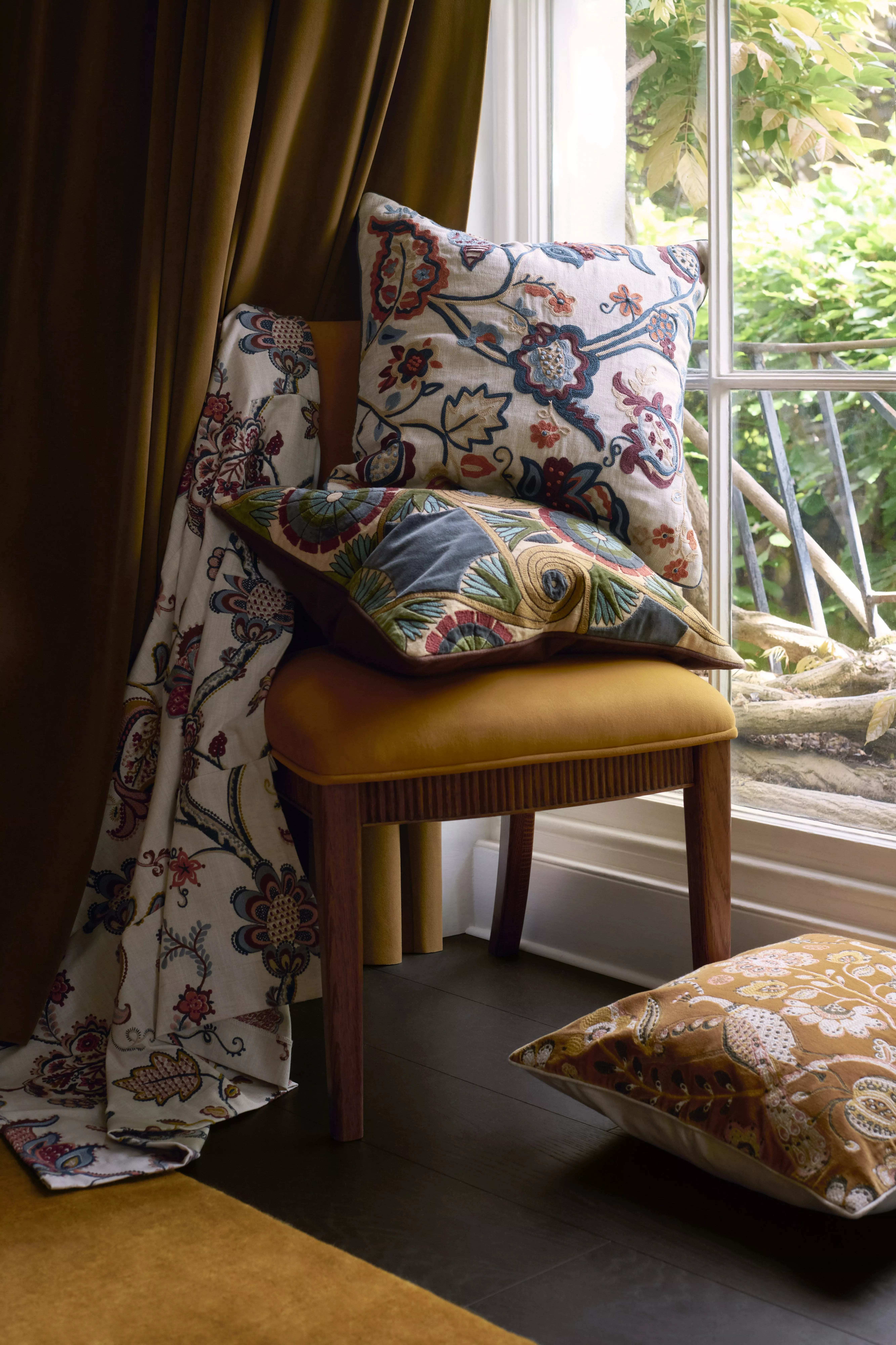 Patterned cushions on a mustard chair by a window, with floral fabric and green garden view beyond