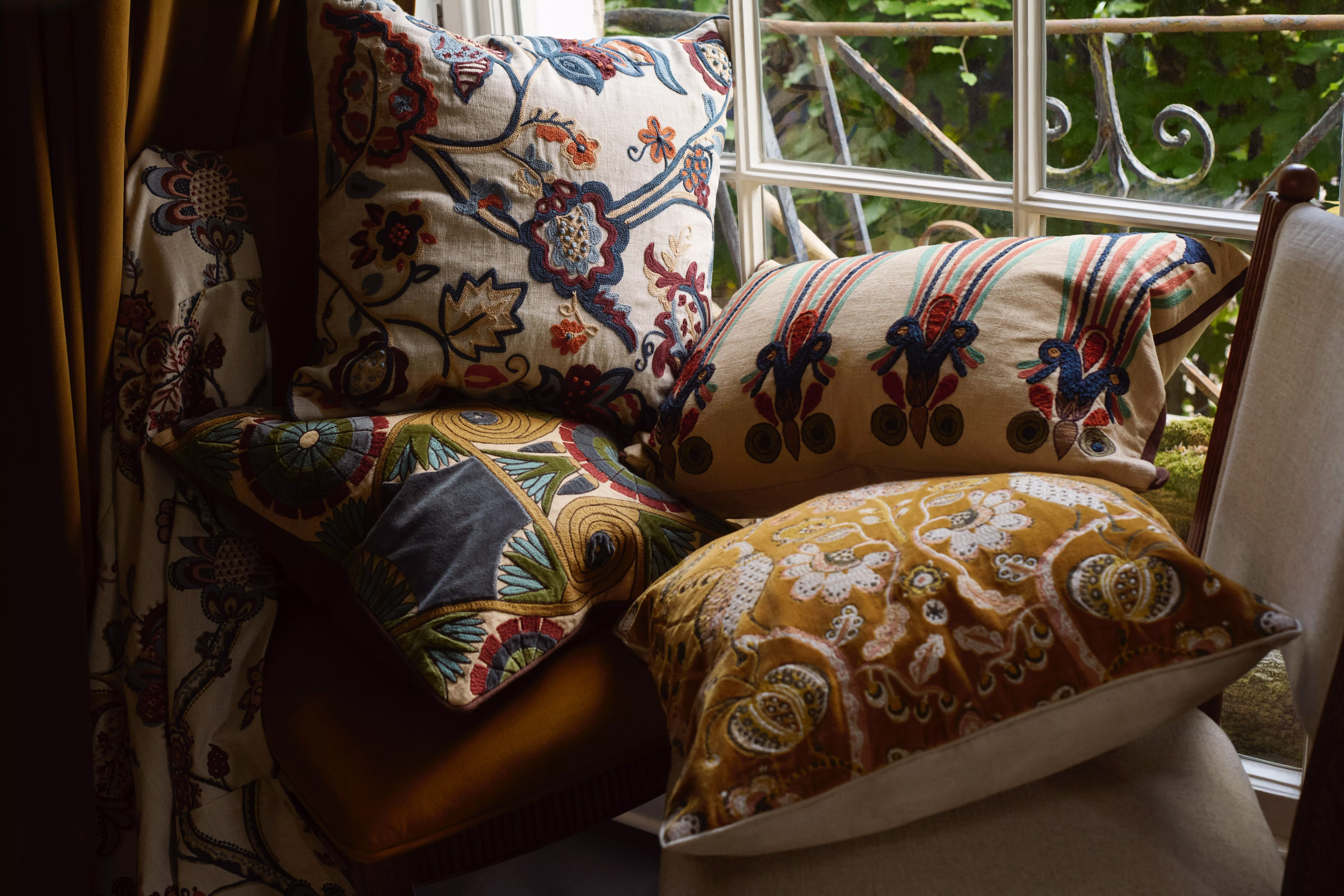 Embroidered patterned cushions on an armchair by window, cosy interior décor with floral and striped designs.