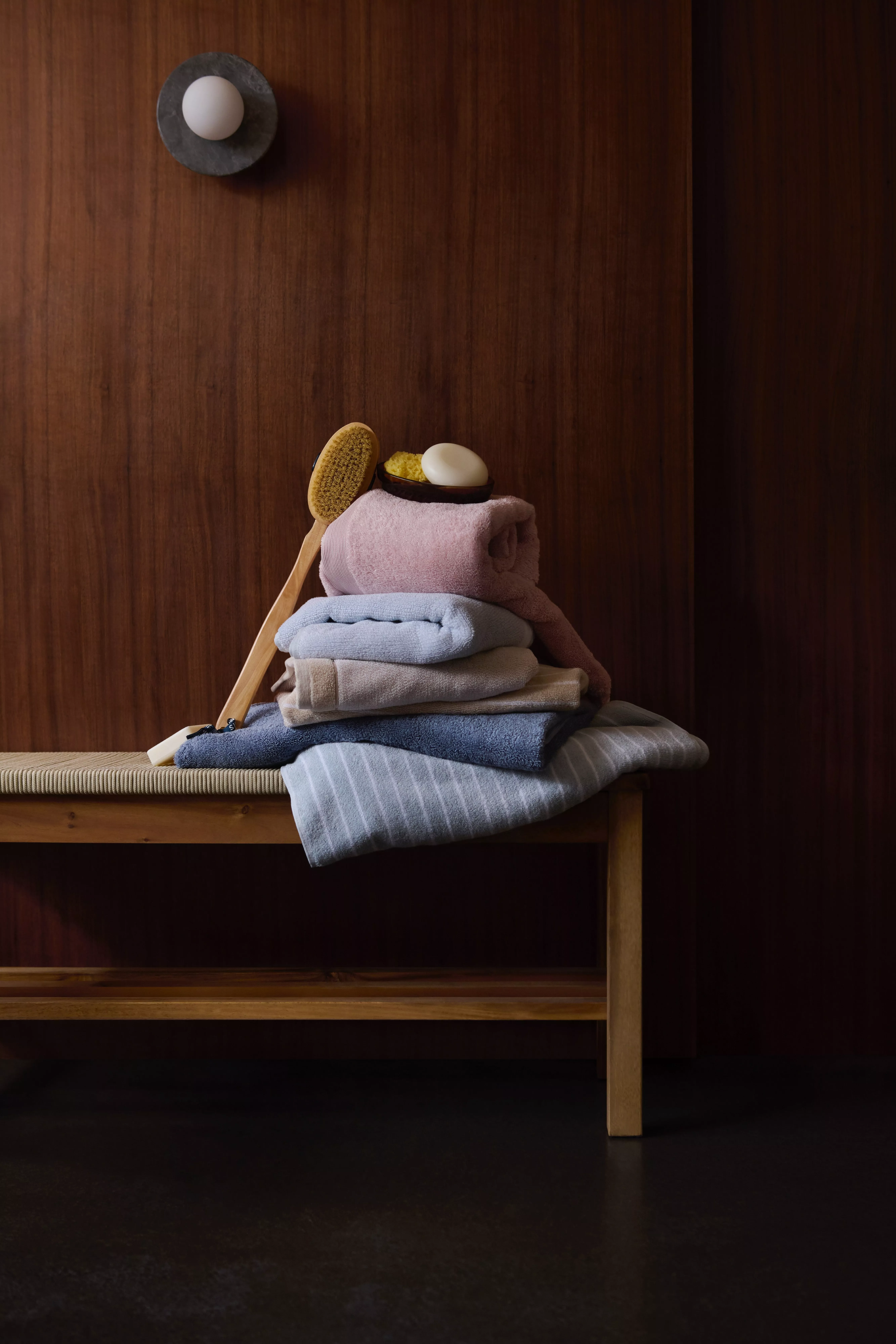 Stack of folded towels with bath brush and soap on a wooden bench, spa bathroom essentials on wood wall backdrop