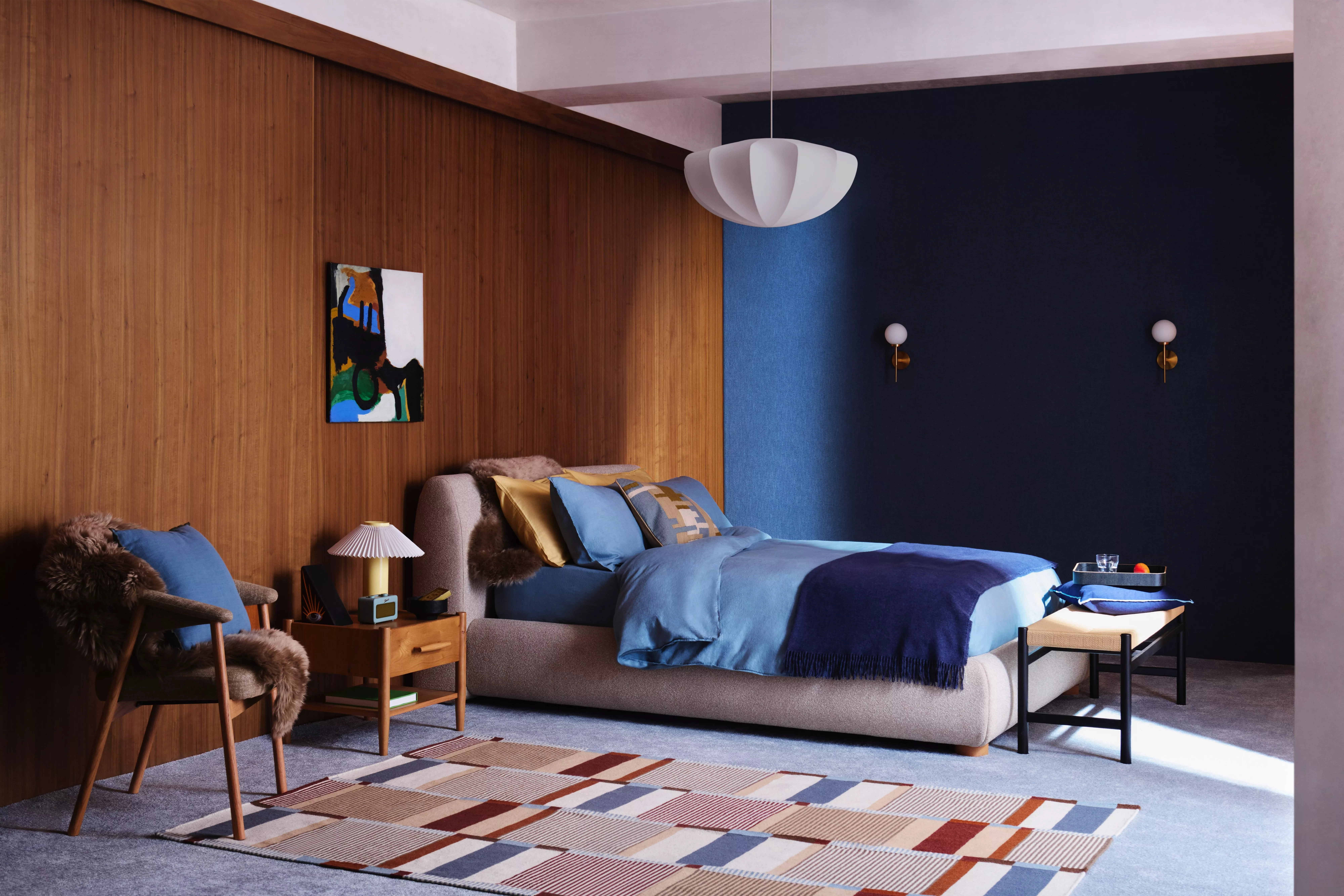 Modern bedroom interior with blue bedding, wood-panel wall, pendant light and minimalist bedside furniture.