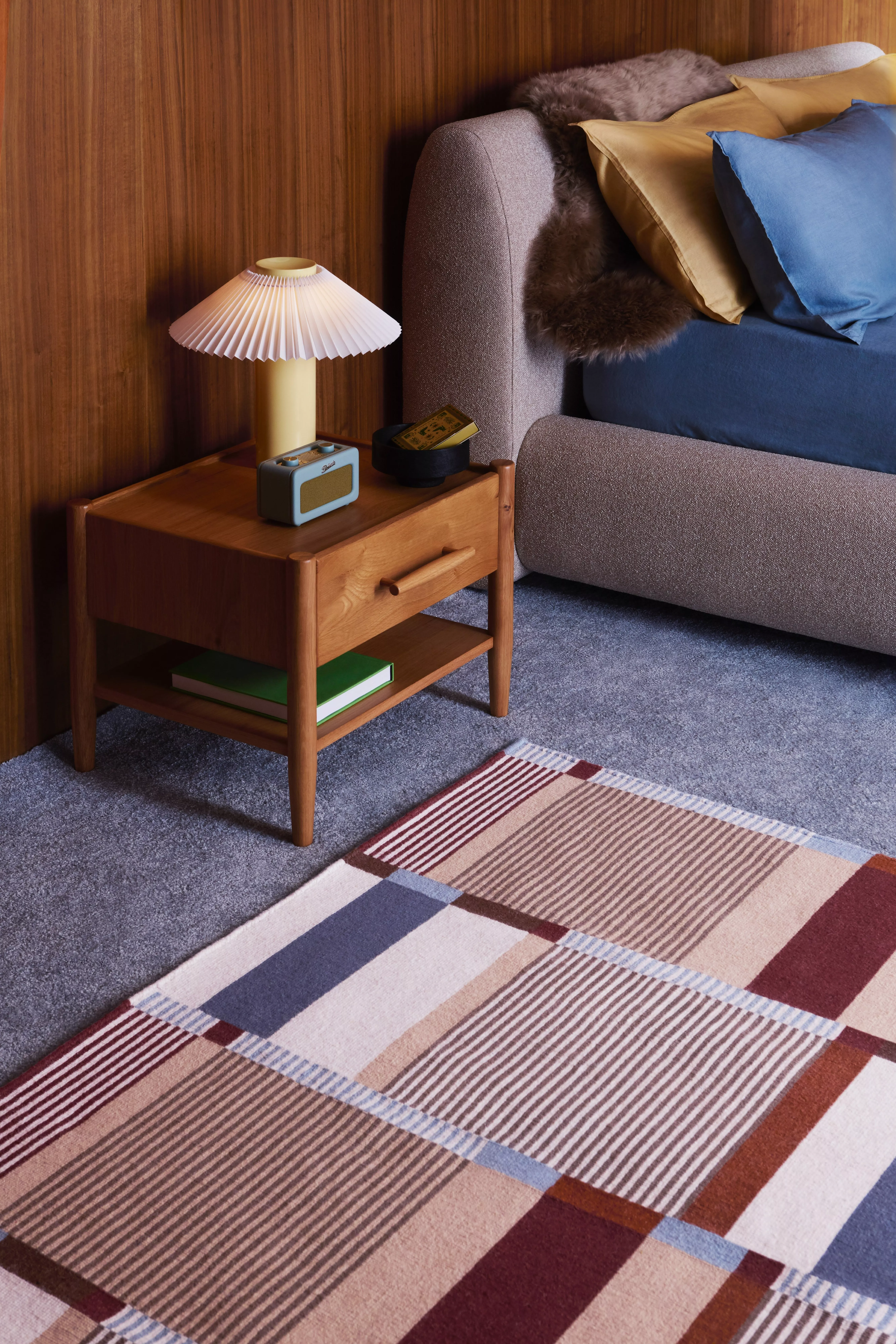Mid-century bedroom corner with wooden bedside table, pleated lamp, radio, and grey bed with cushions and rug