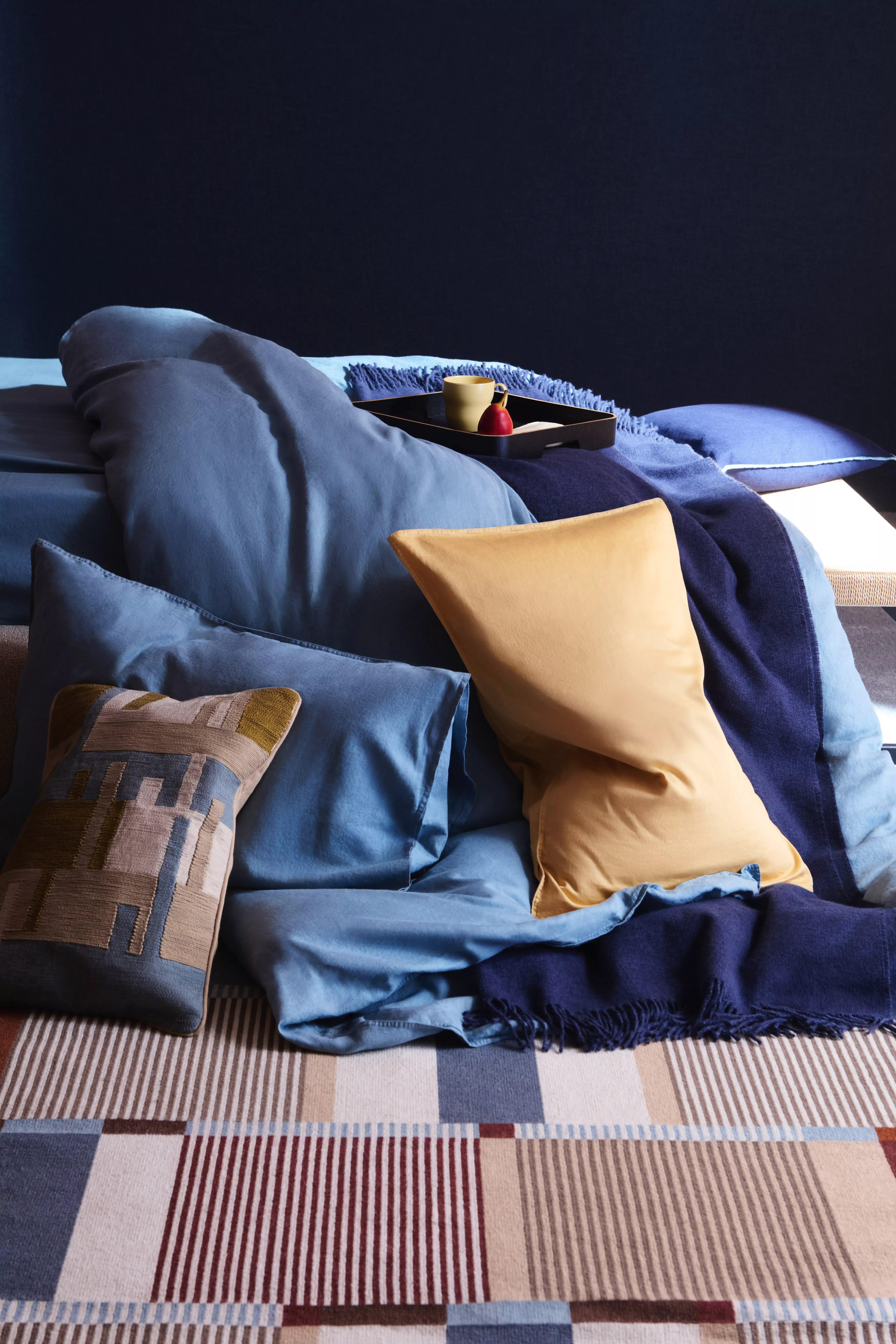 Modern bedroom with blue bedding, navy throw and mustard cushions, tray with mug on striped bedspread