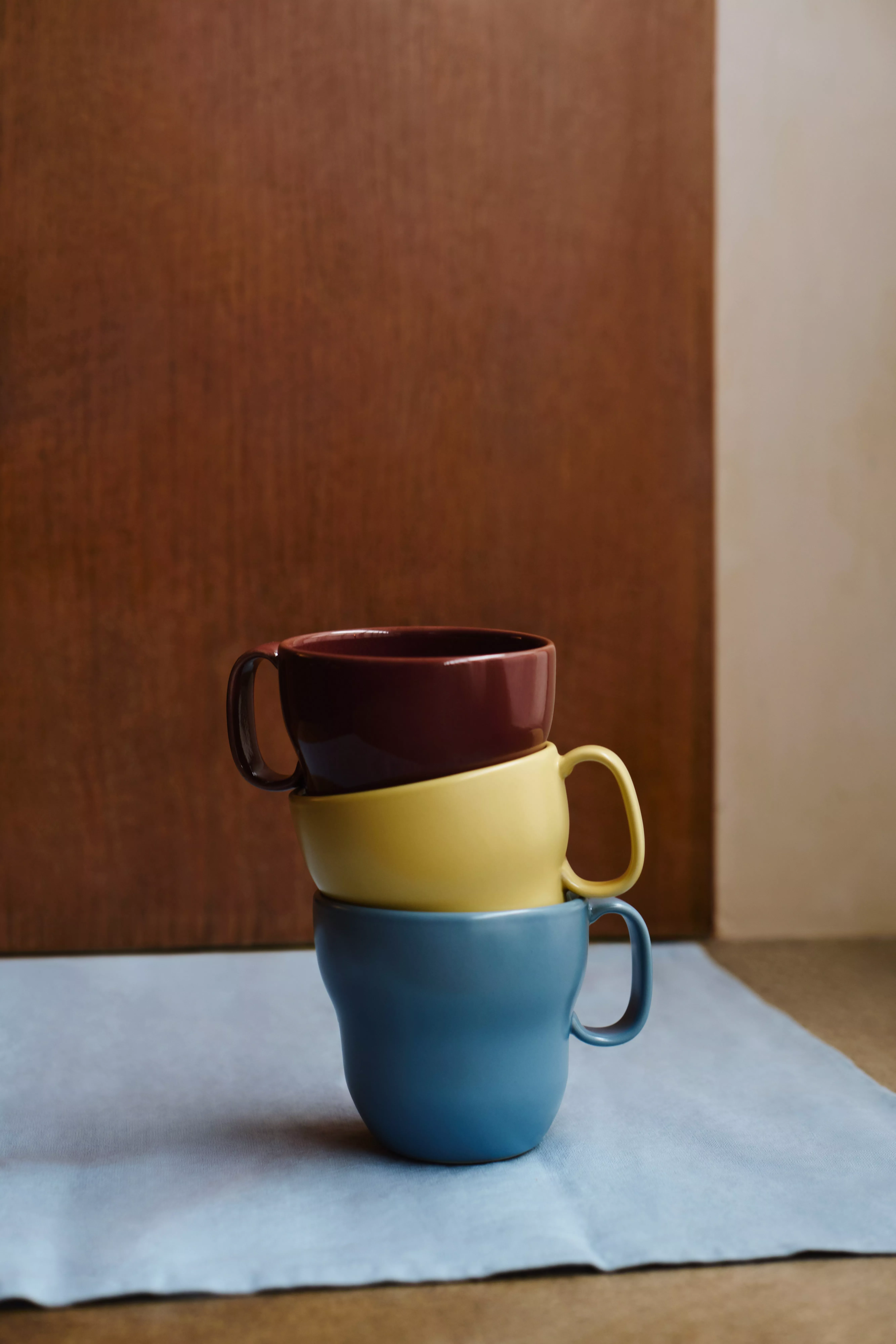 Stacked colourful ceramic coffee mugs on a blue cloth, minimalist kitchen decor against wooden background