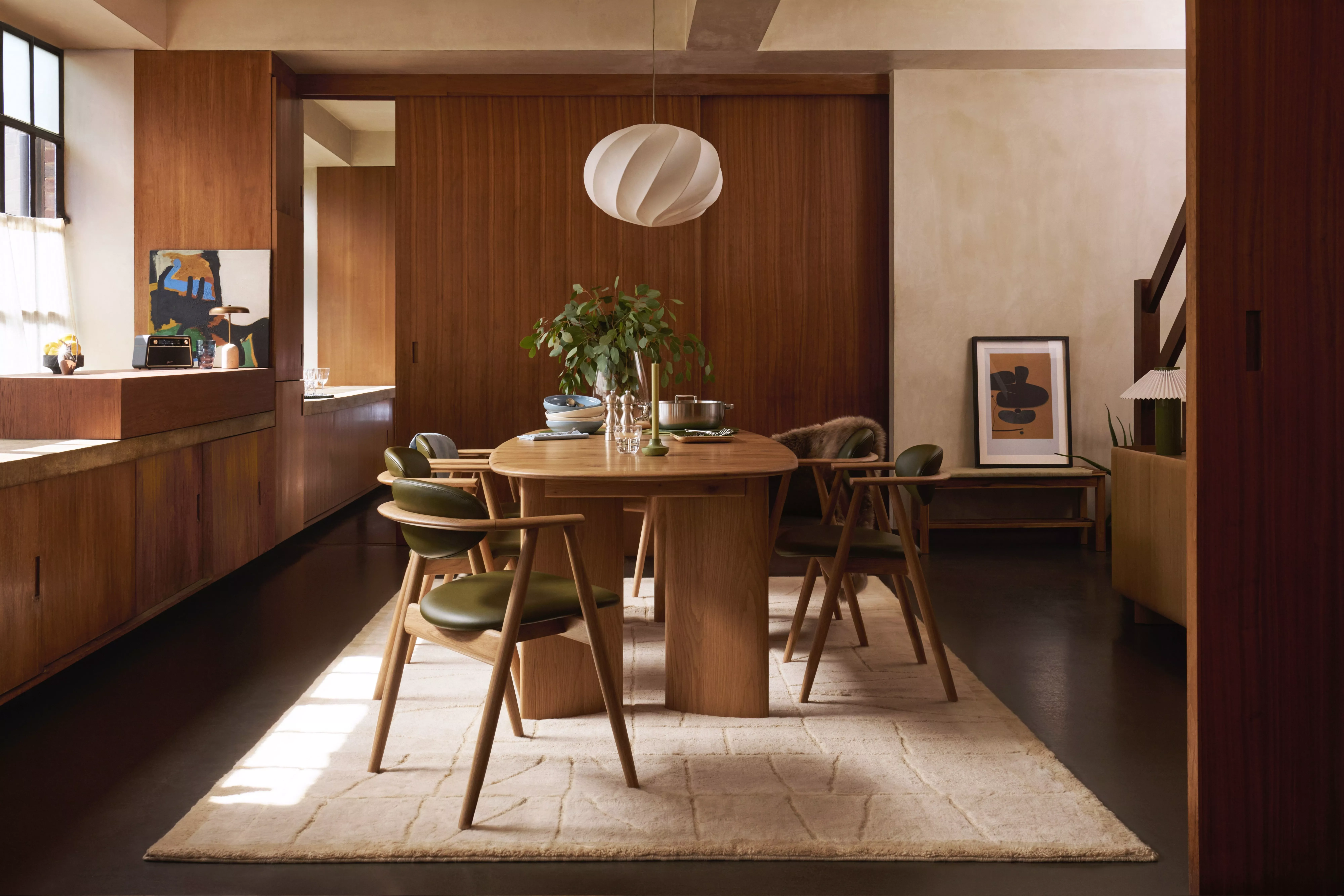 Mid-century modern dining room with wood panelling, oak table, green chairs, pendant light and artwork.