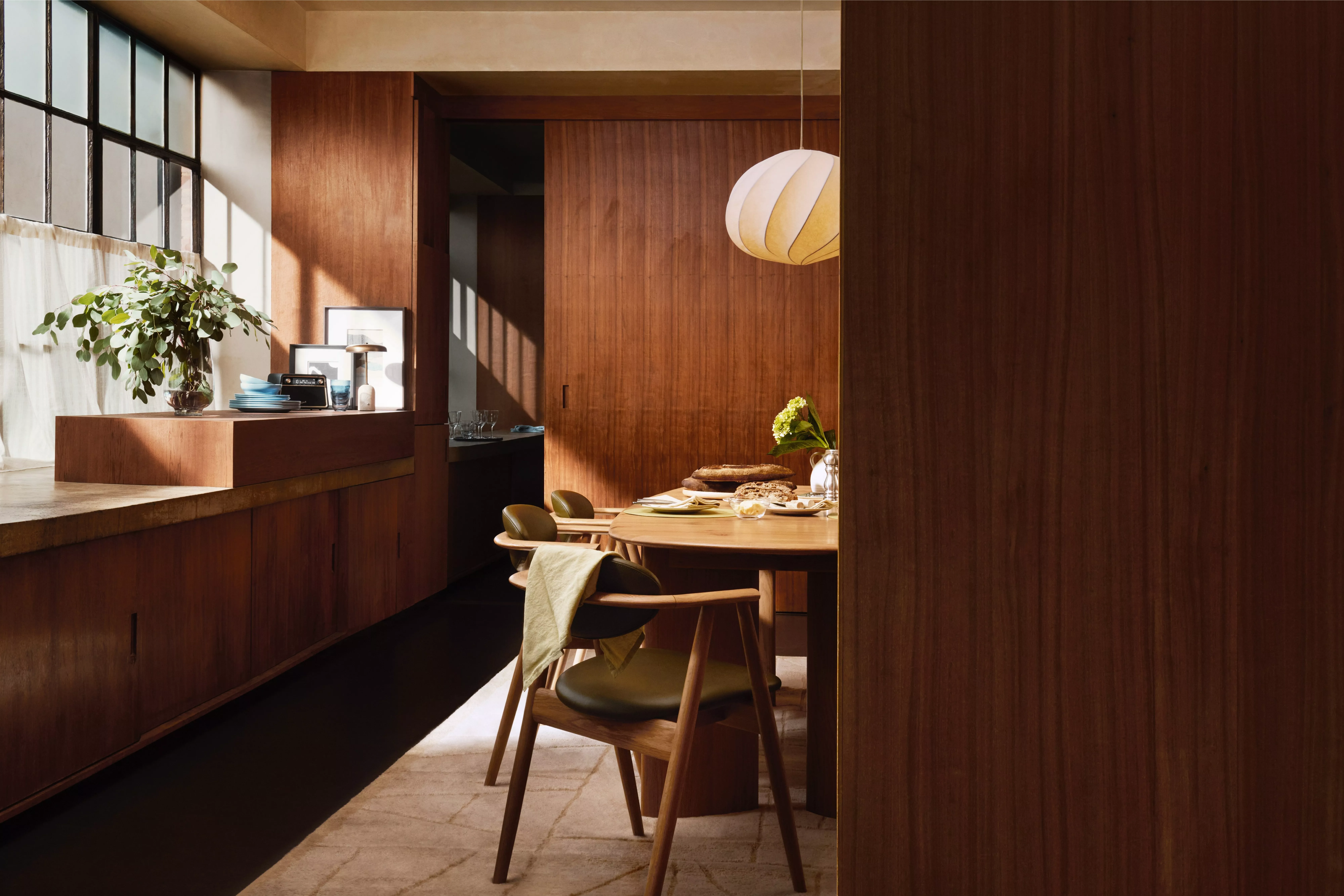 Sunlit modern dining room with warm wood panelling, round table, chairs, pendant lamp and plant by window