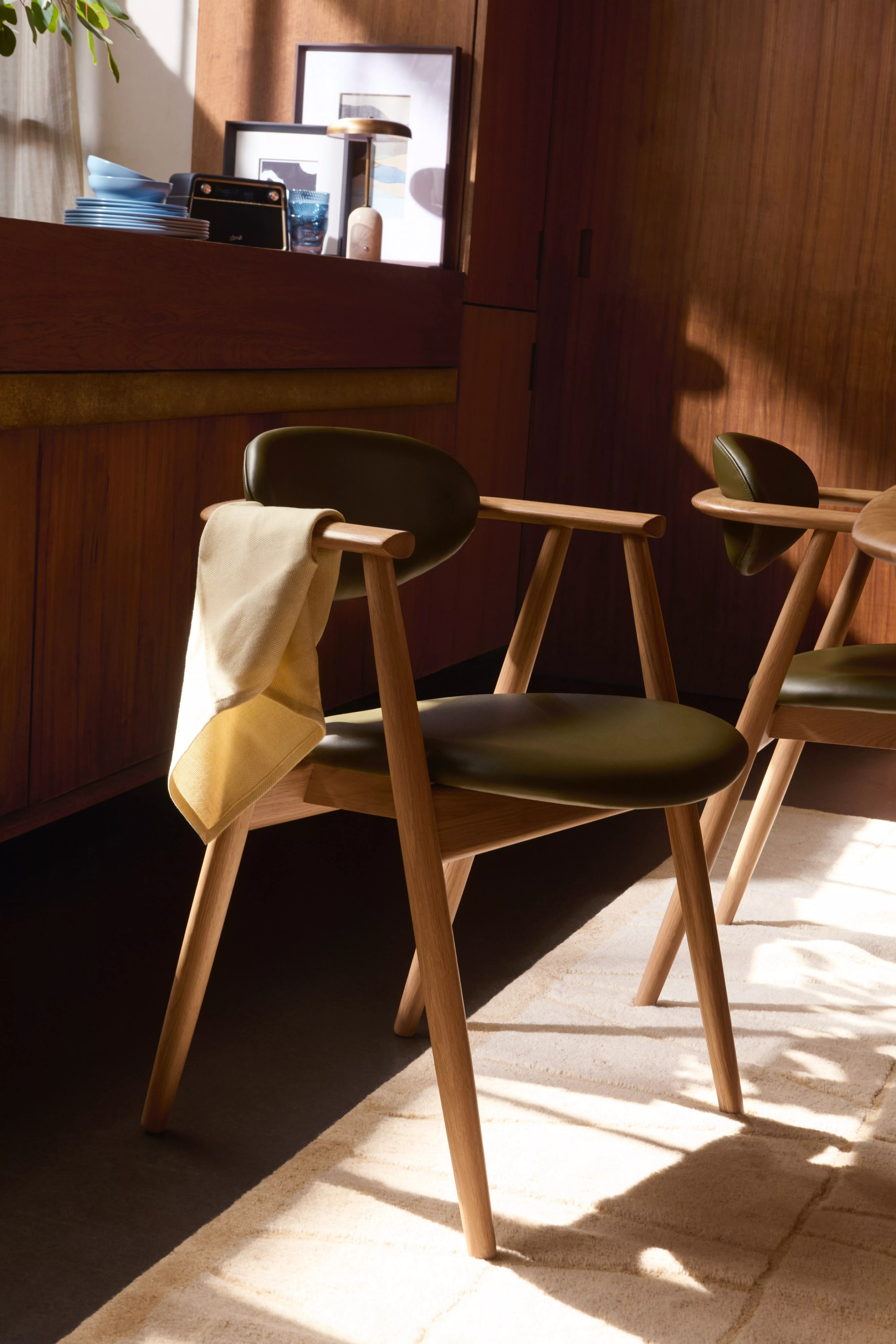 Sunlit modern dining chairs with green upholstery and oak arms in a warm wooden interior setting