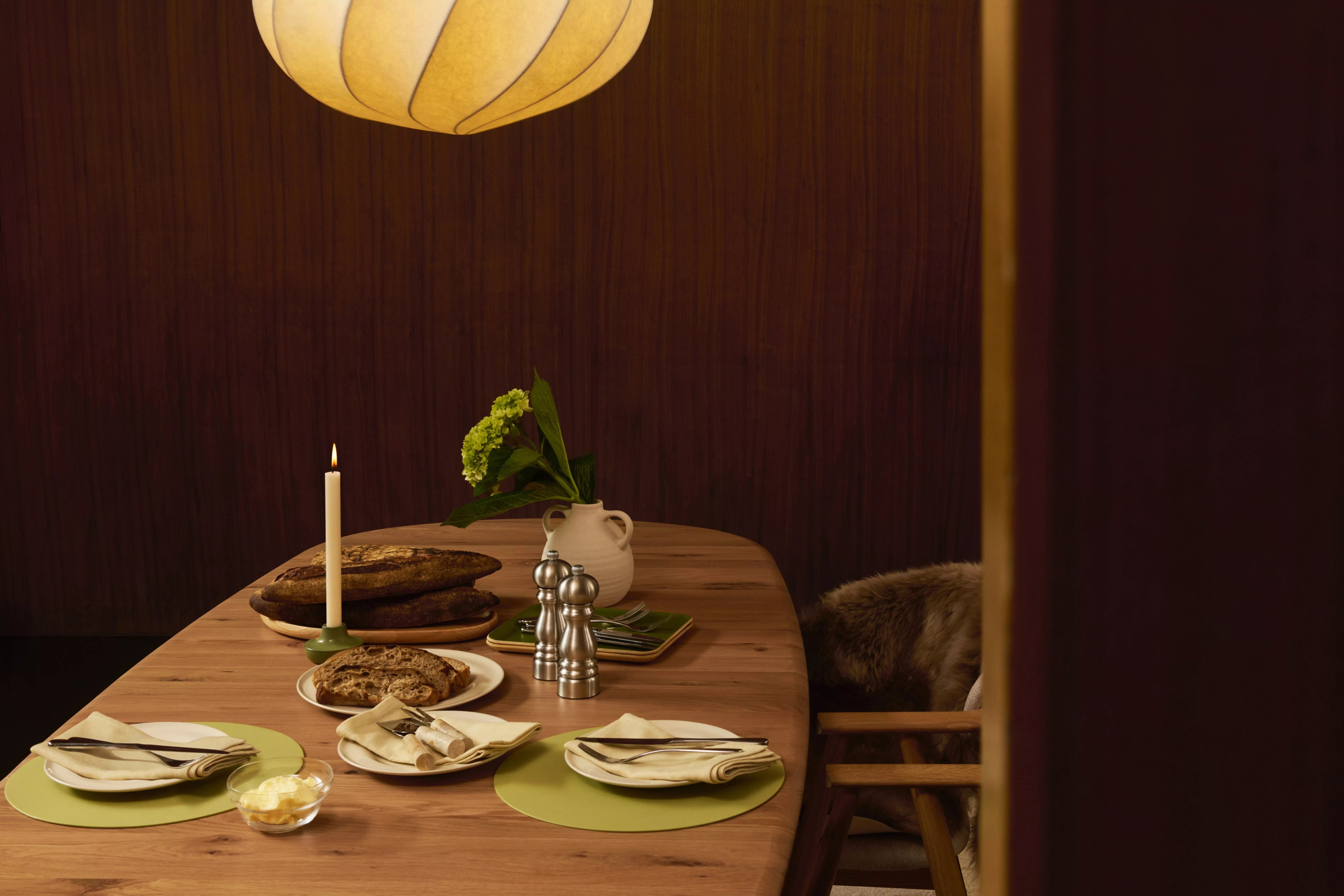 Cosy dining table set with bread, candle, cutlery and vase of flowers under warm pendant light in wooden room