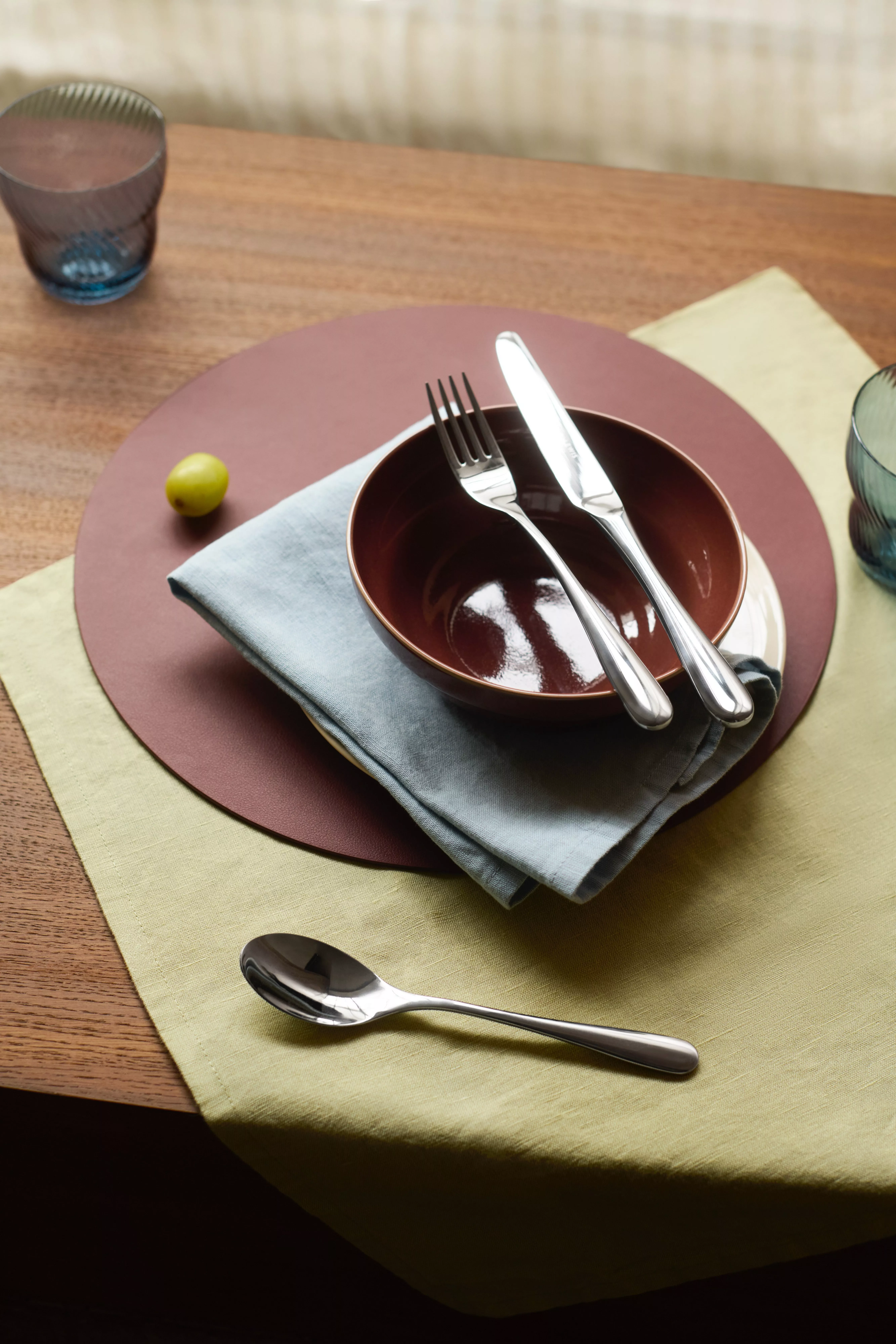 Modern table setting with ceramic bowl, fork and knife on napkin, spoon, placemat and tumblers on wooden table.