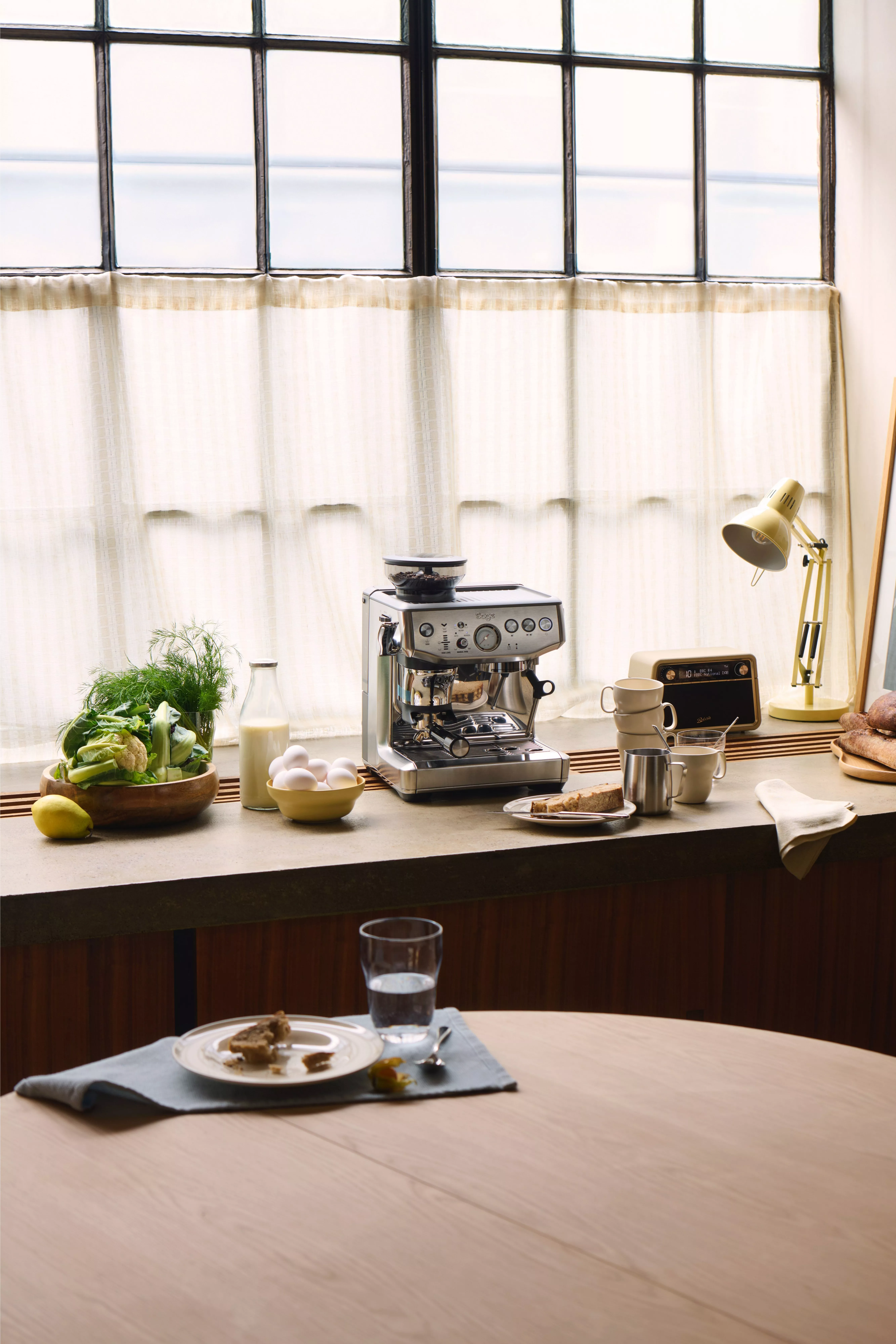 Sunlit kitchen counter with espresso machine, kettle, mugs, milk and fresh vegetables beside framed wall art