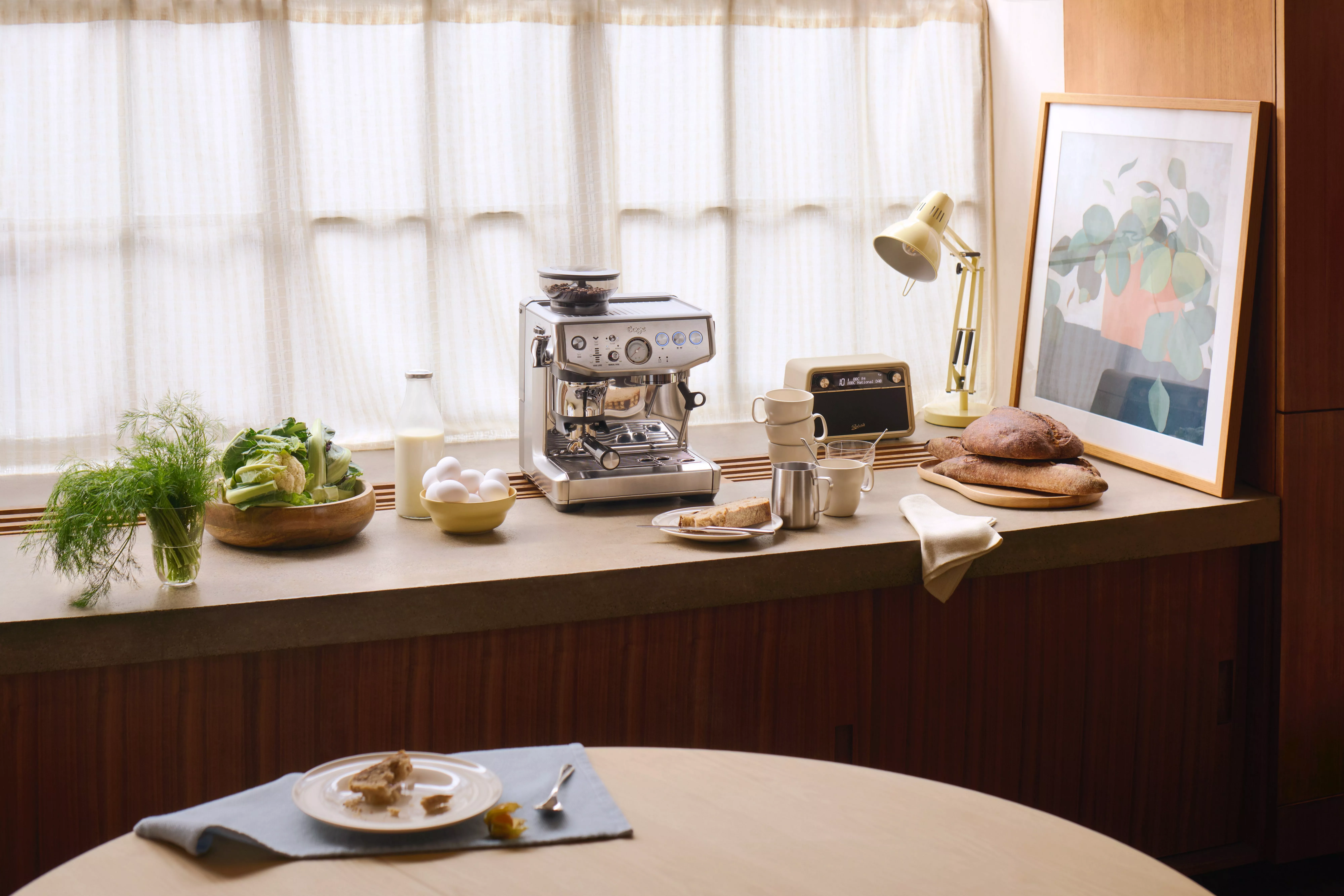 Sunlit kitchen counter with espresso machine, fresh bread, eggs, milk and herbs beside a framed print and lamp