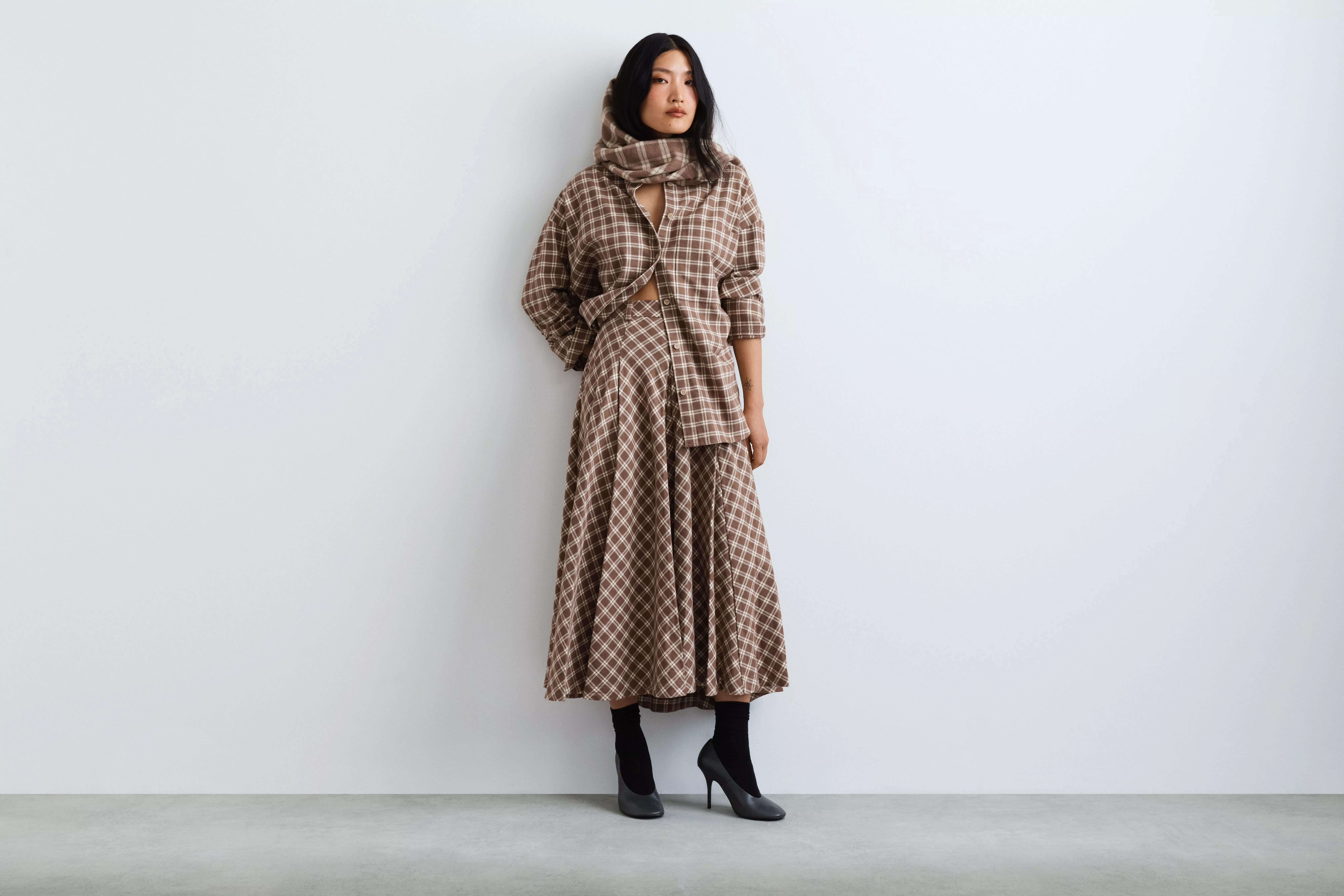 Fashion model wearing brown checked co-ord skirt and jacket with scarf, posing against a minimalist white wall