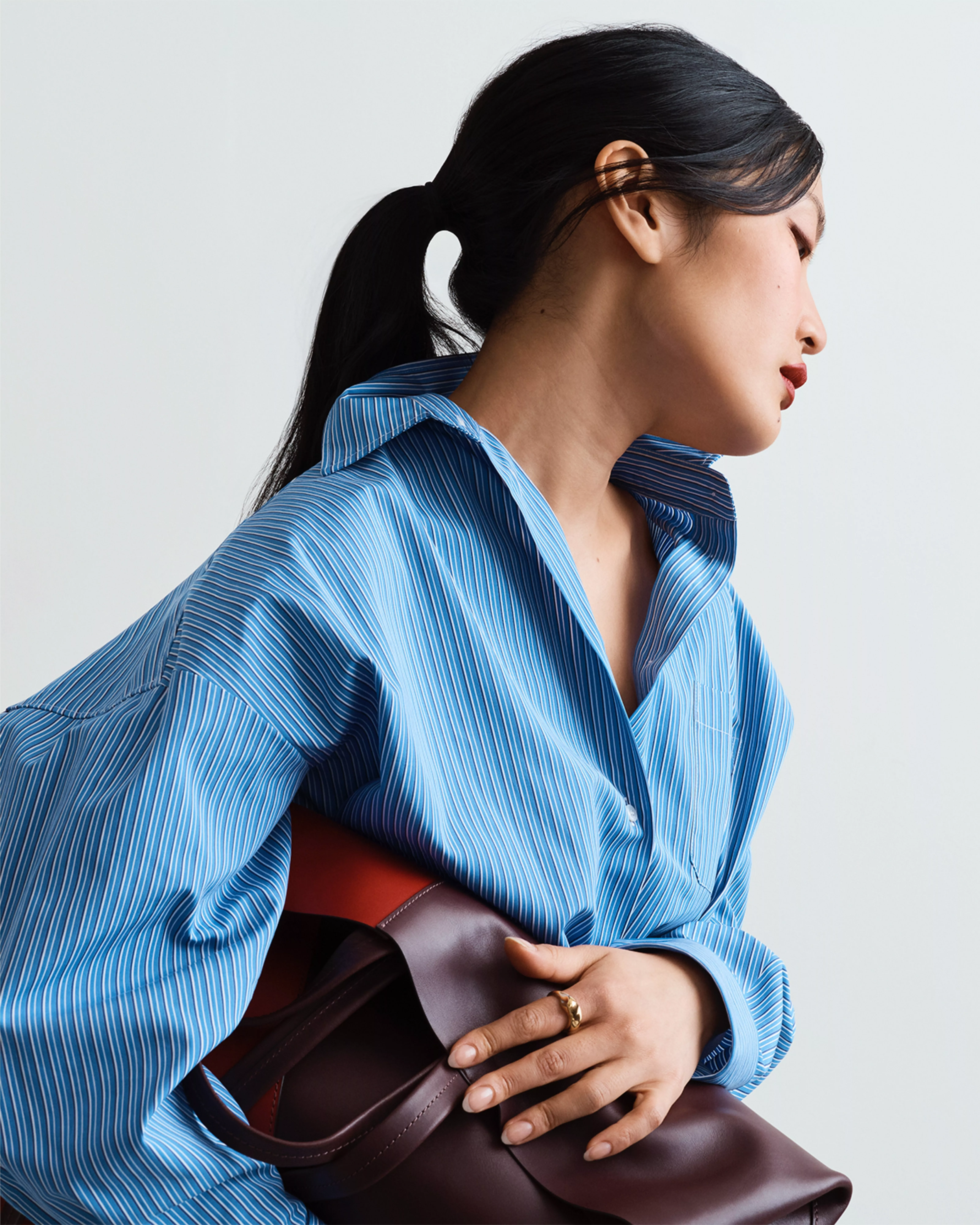 Side profile of woman in blue striped shirt holding a brown leather handbag, minimalist fashion portrait on grey background