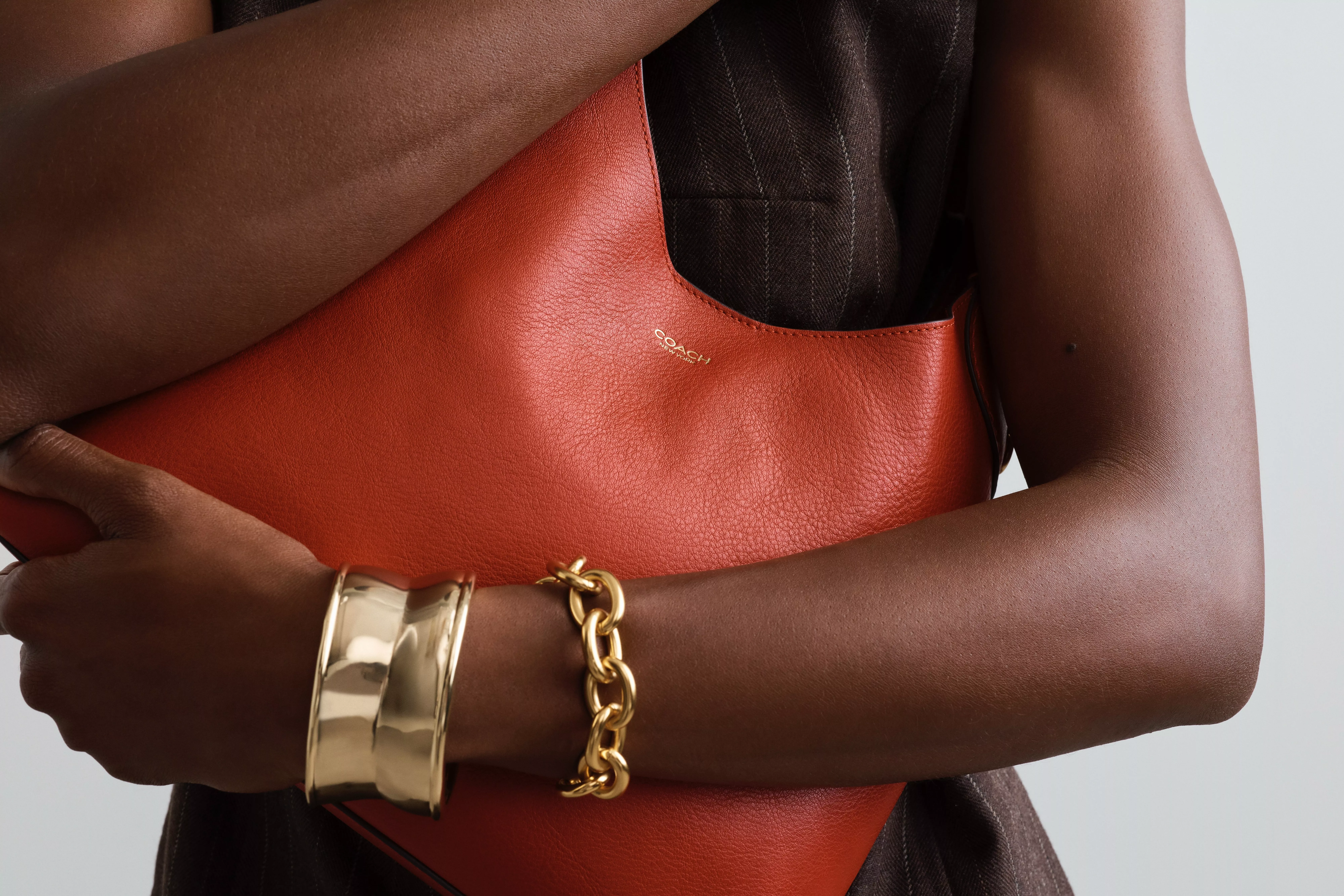 Close-up of model holding a burnt orange Coach leather shoulder bag, wearing gold bangle and chain bracelet.
