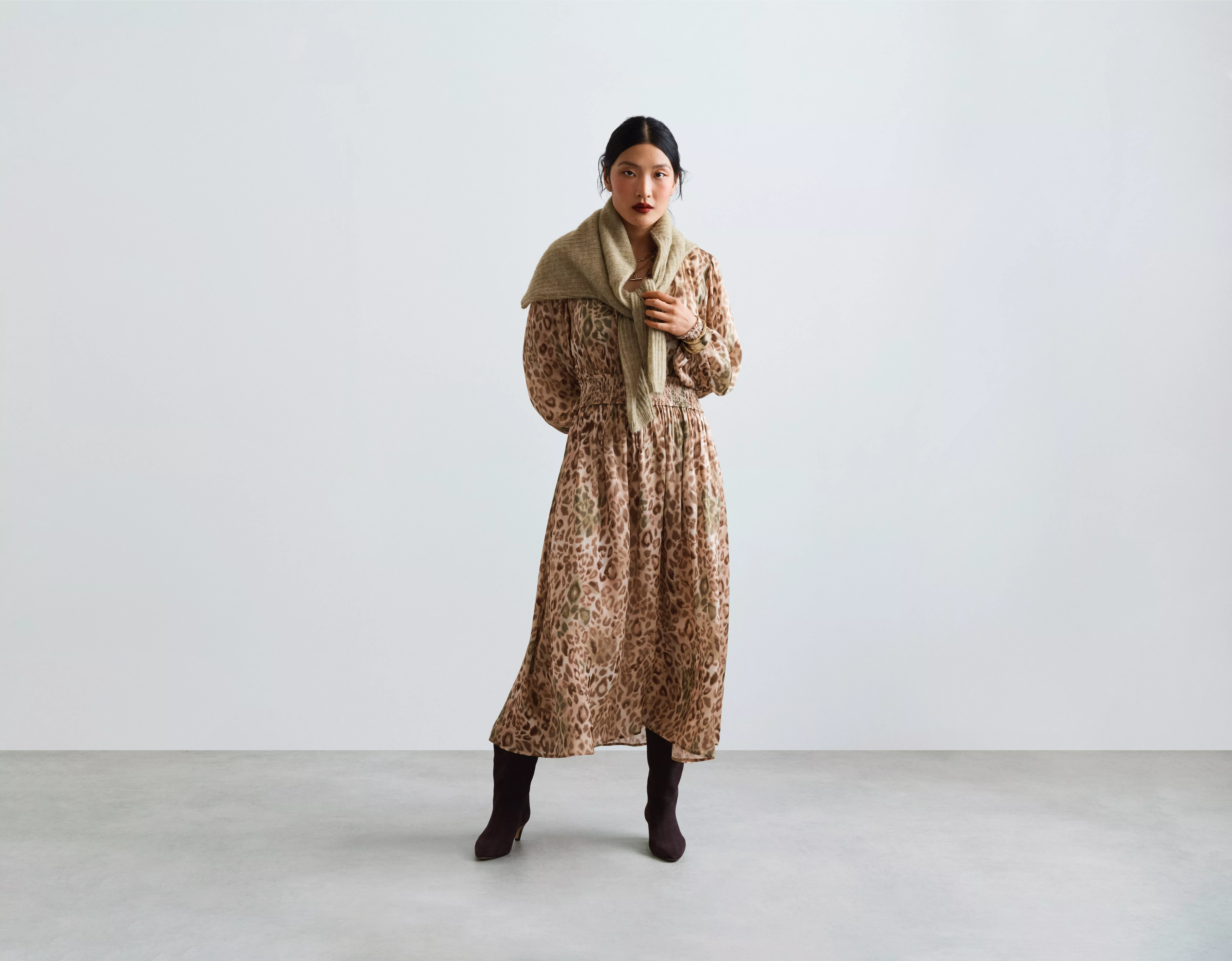 Full-length studio fashion portrait of woman in leopard-print midi dress, beige scarf and black boots on grey backdrop