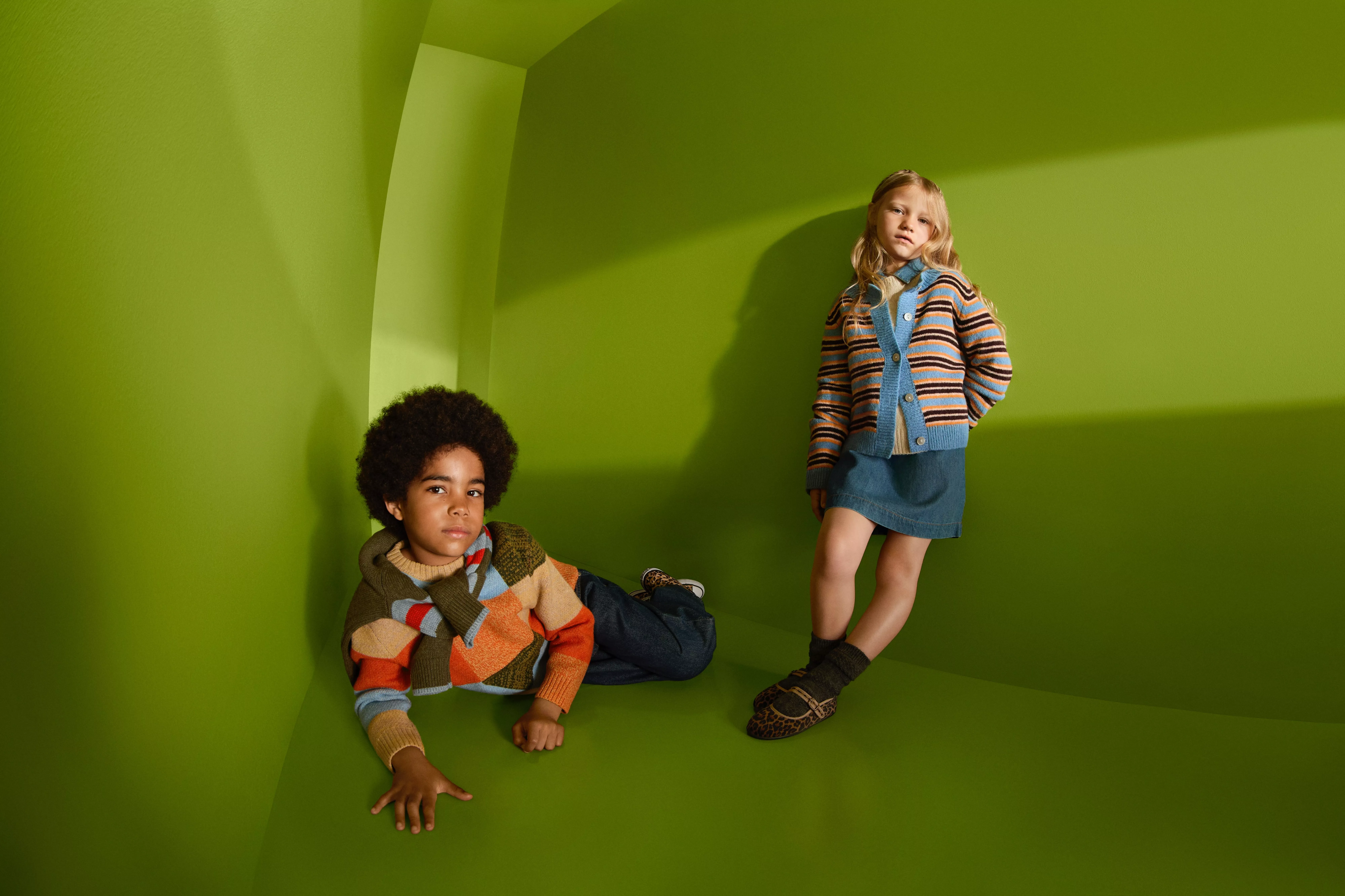 Two children modelling autumn knitwear and denim outfits in a green studio corner, relaxed fashion portrait