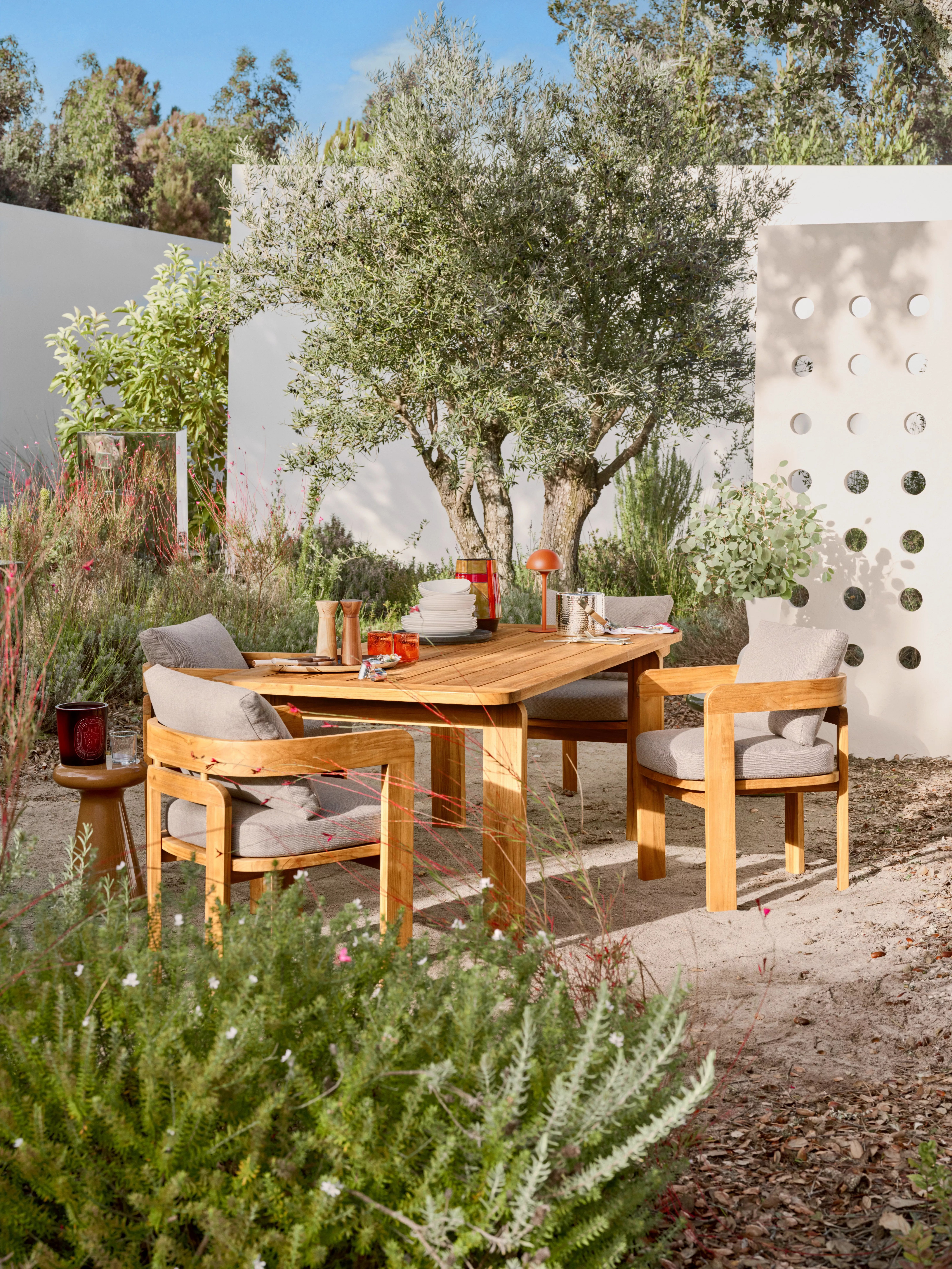 Outdoor dining area with wooden furniture, surrounded by lush greenery and a modern, white perforated wall.