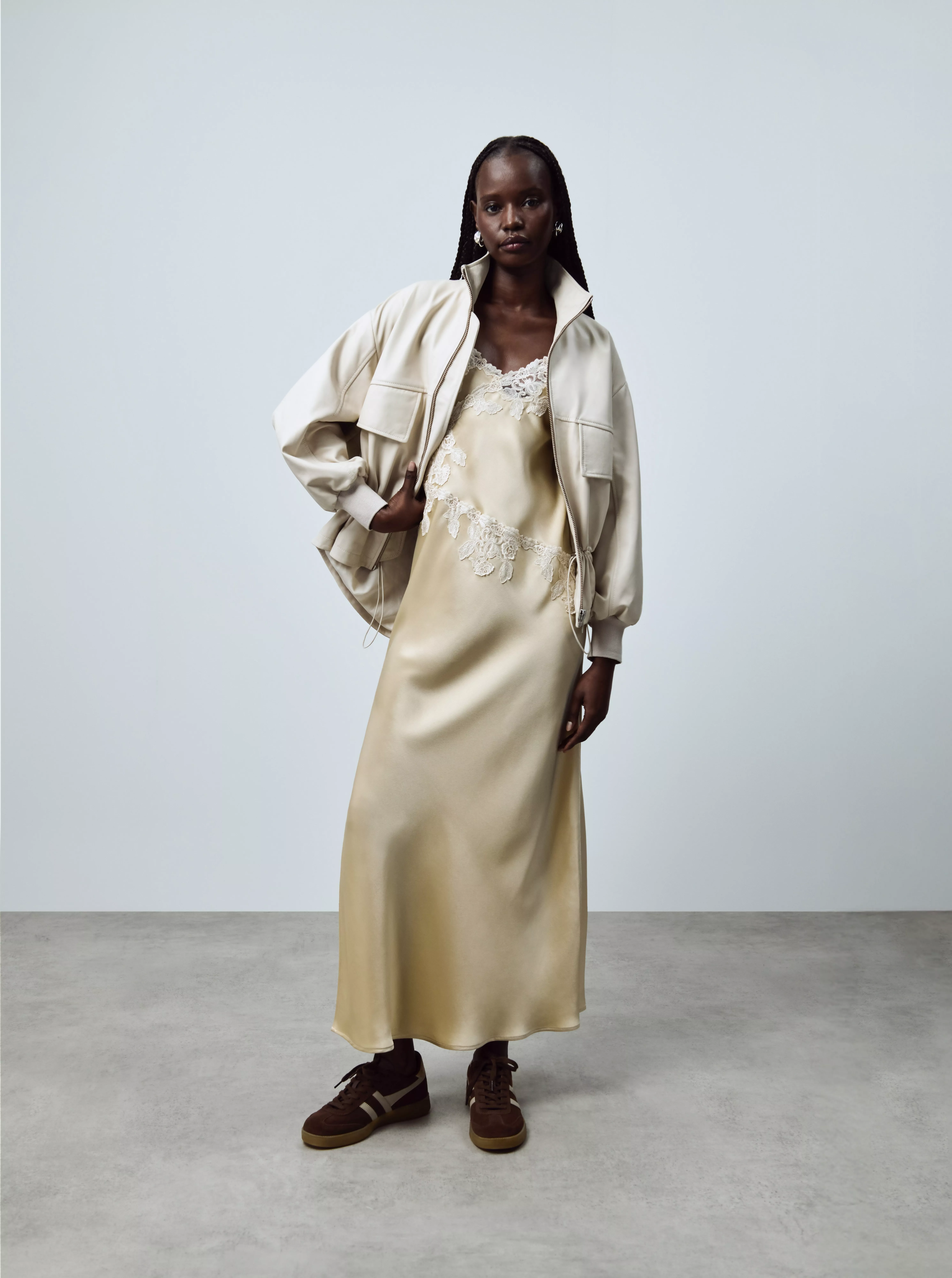 Model in satin slip dress with lace trim and cream bomber jacket, styled with brown trainers in studio setting