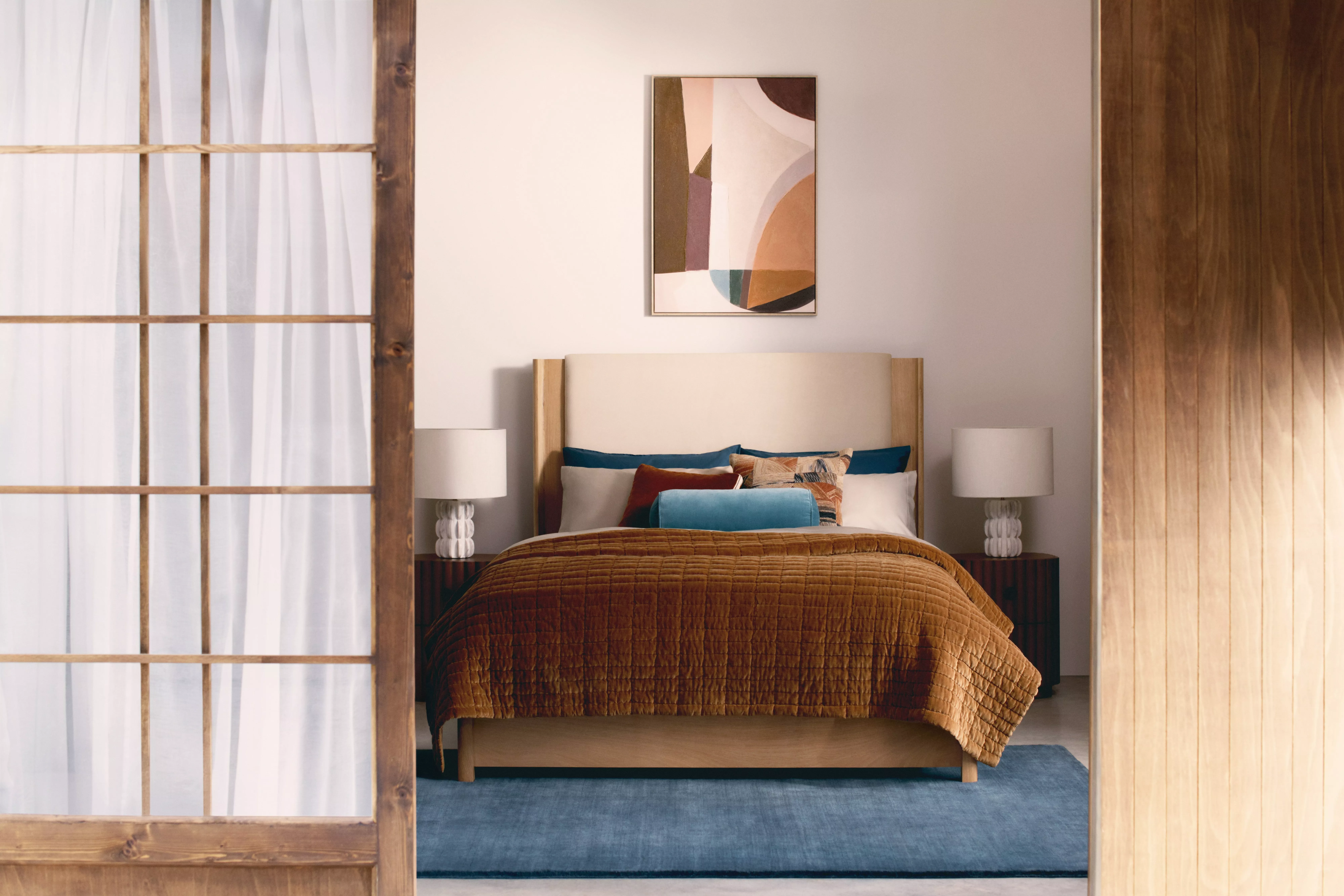 Cosy bedroom with wooden sliding doors, abstract artwork, plush bedding, and modern lamps on bedside tables.