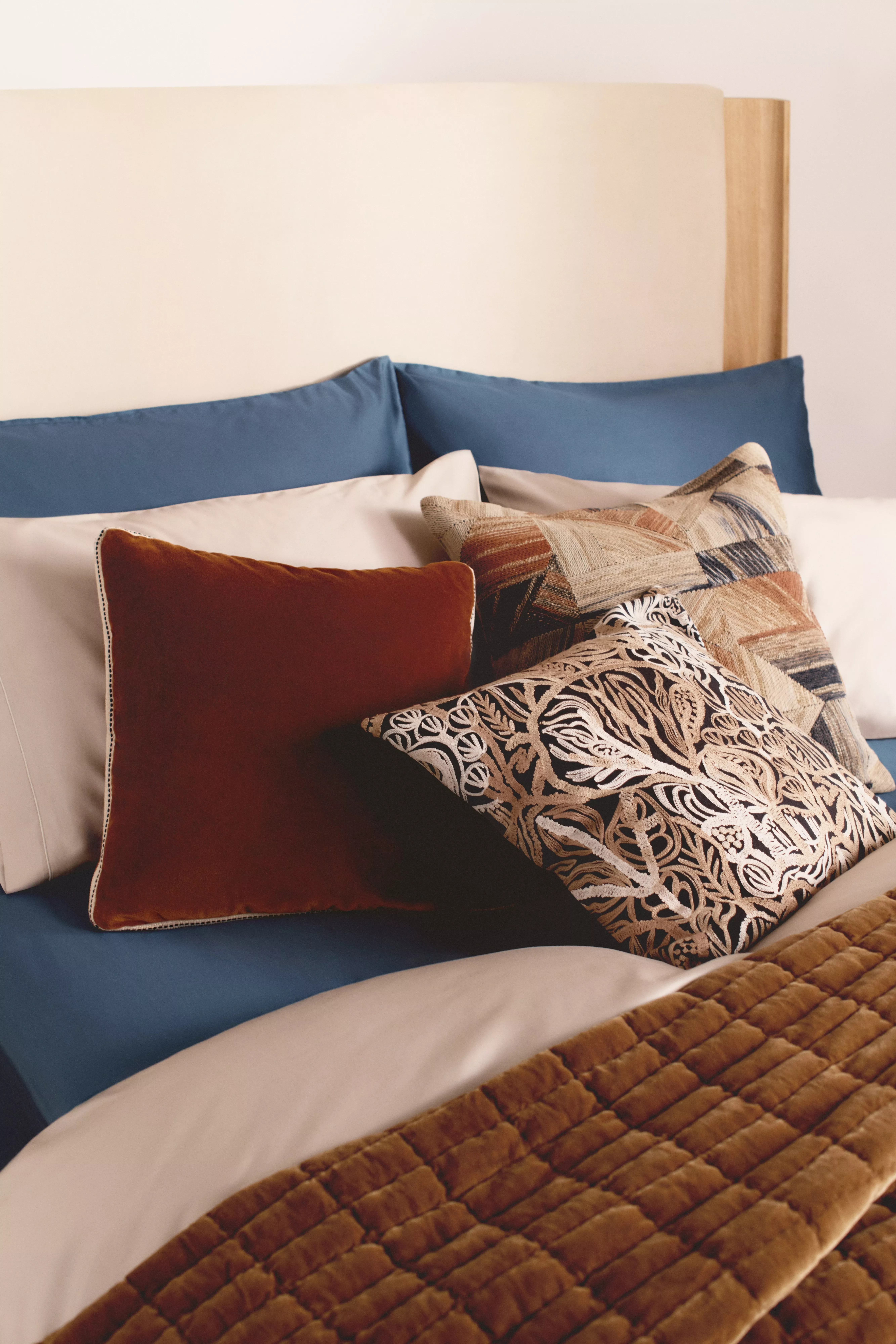 A styled bed showing blue sheets, a rust-colored velvet throw pillow, and a patterned pillow with a brown quilted throw.