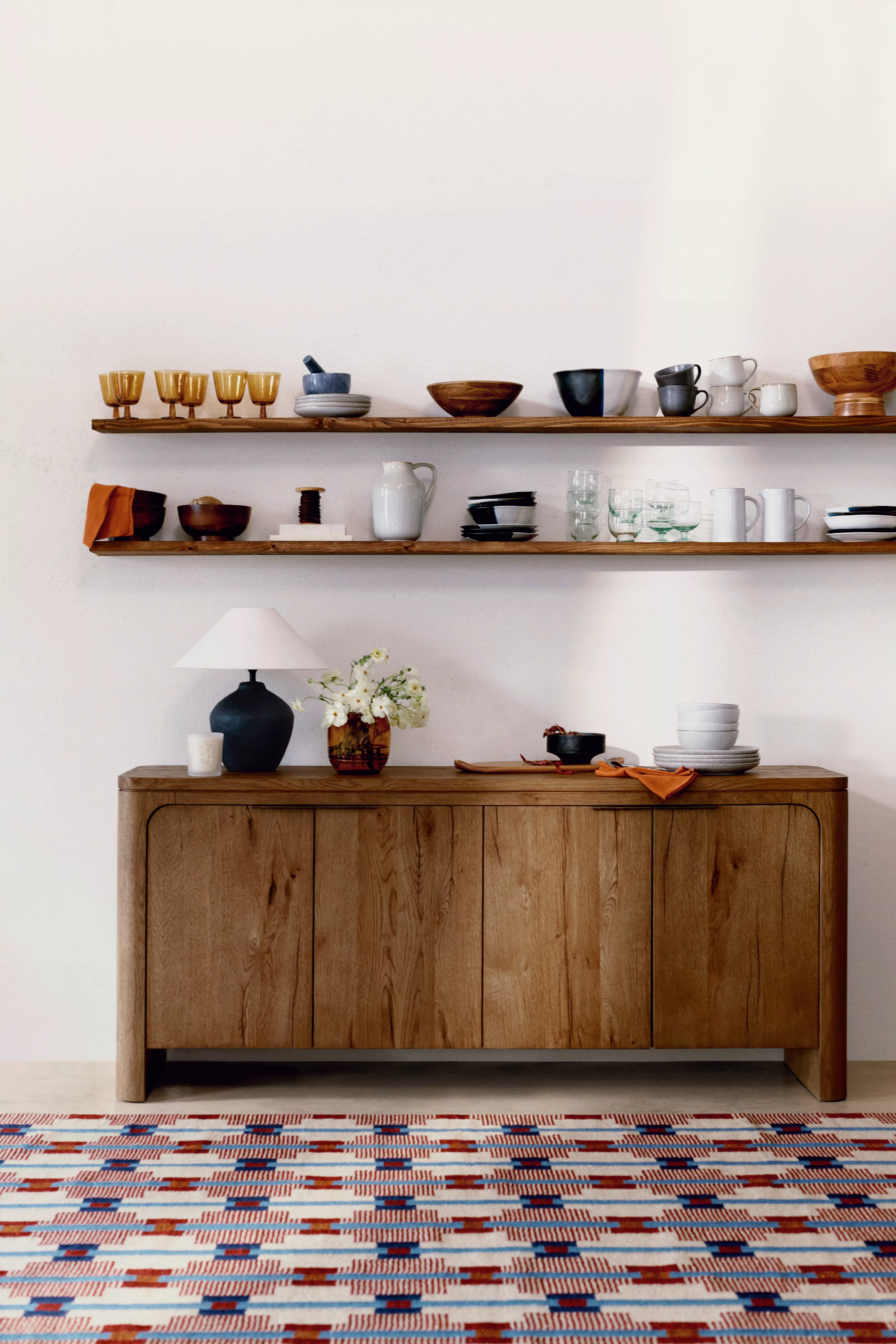 Stylish kitchen shelves with assorted ceramics and glassware above a wooden sideboard on a patterned rug.