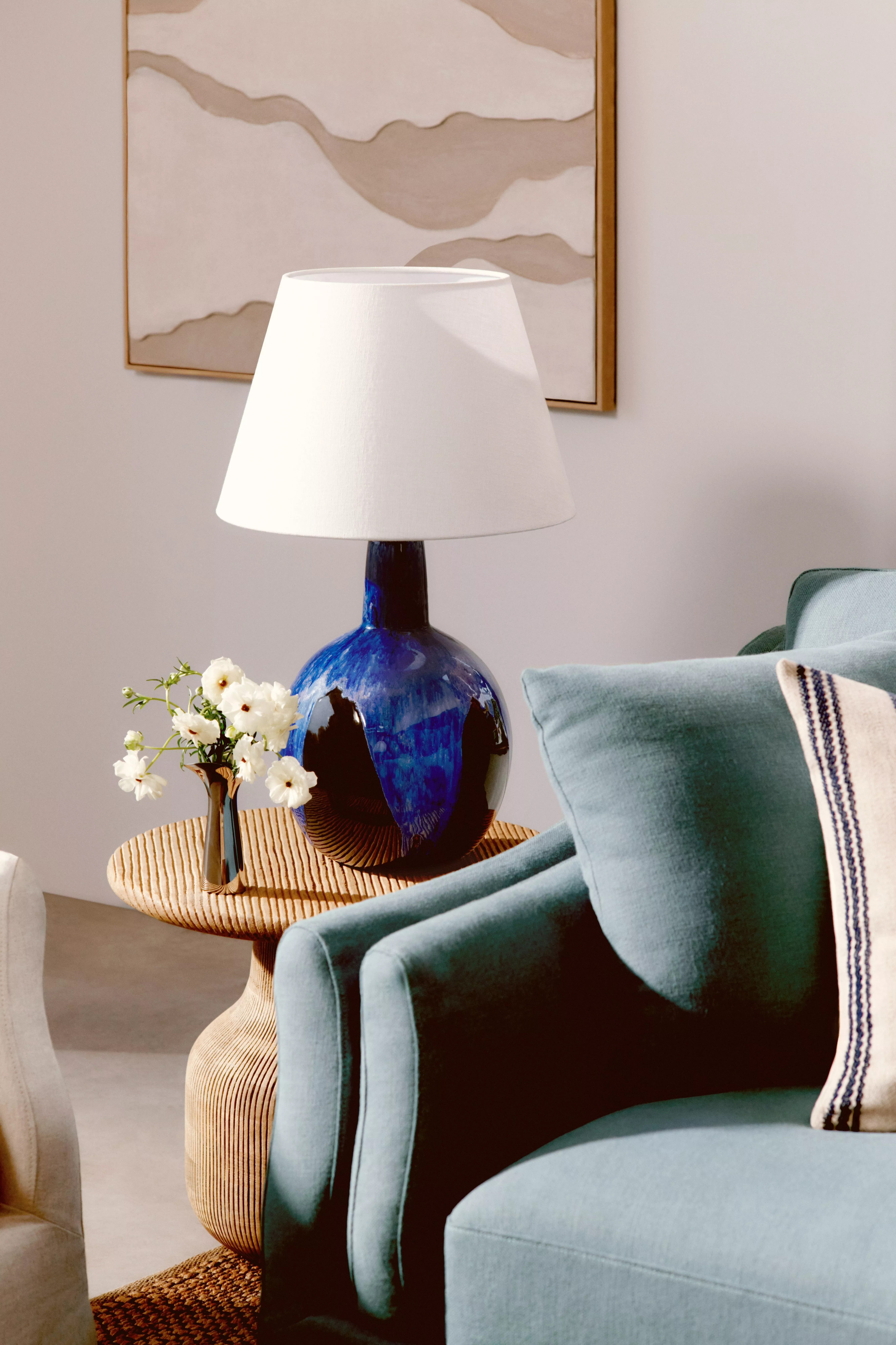 A close-up of a living room corner featuring a glossy cobalt blue table lamp on a woven side table next to a teal armchair.