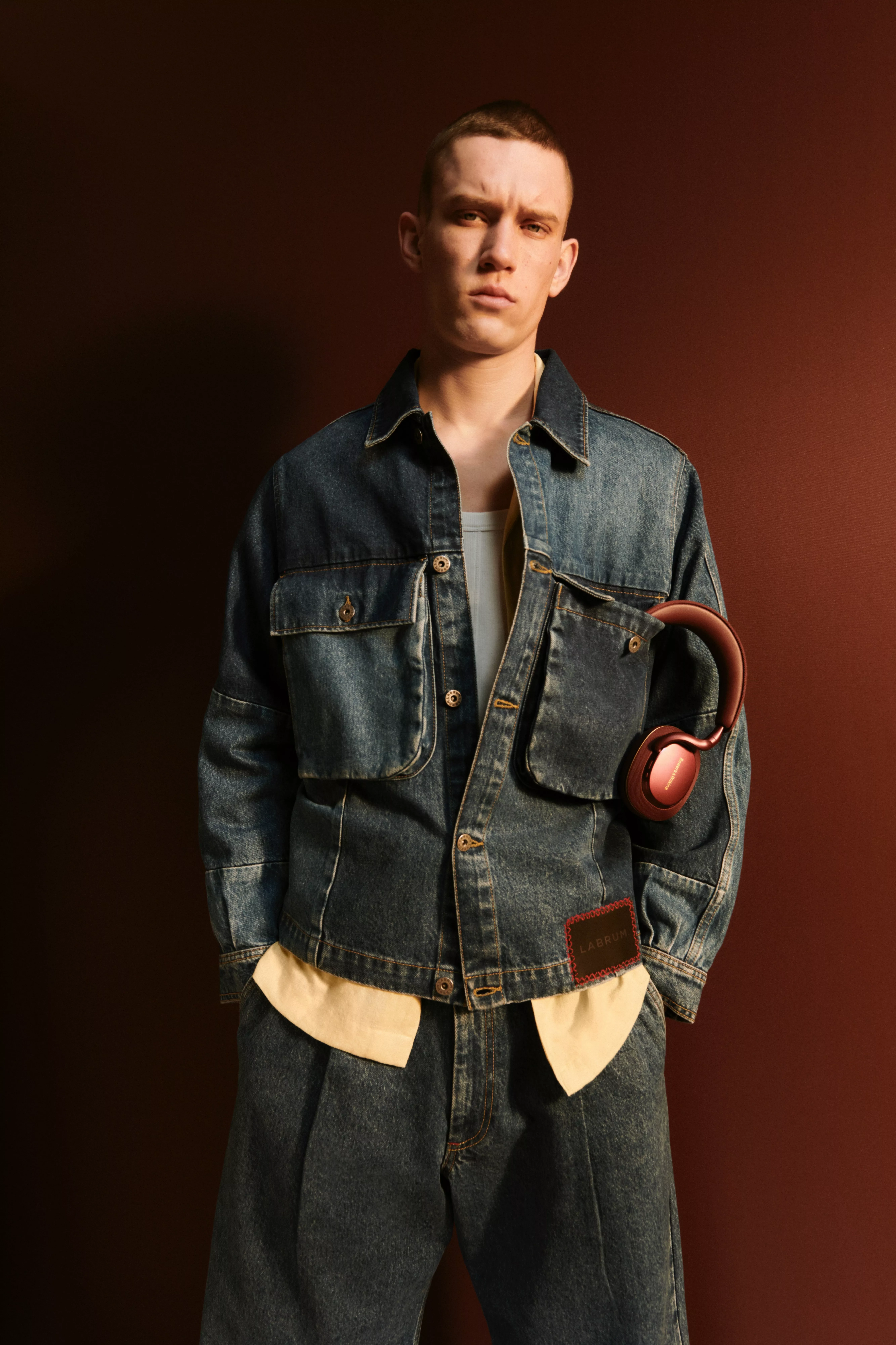 Studio fashion portrait of man in oversized denim jacket and jeans, holding red wireless headphones on dark backdrop