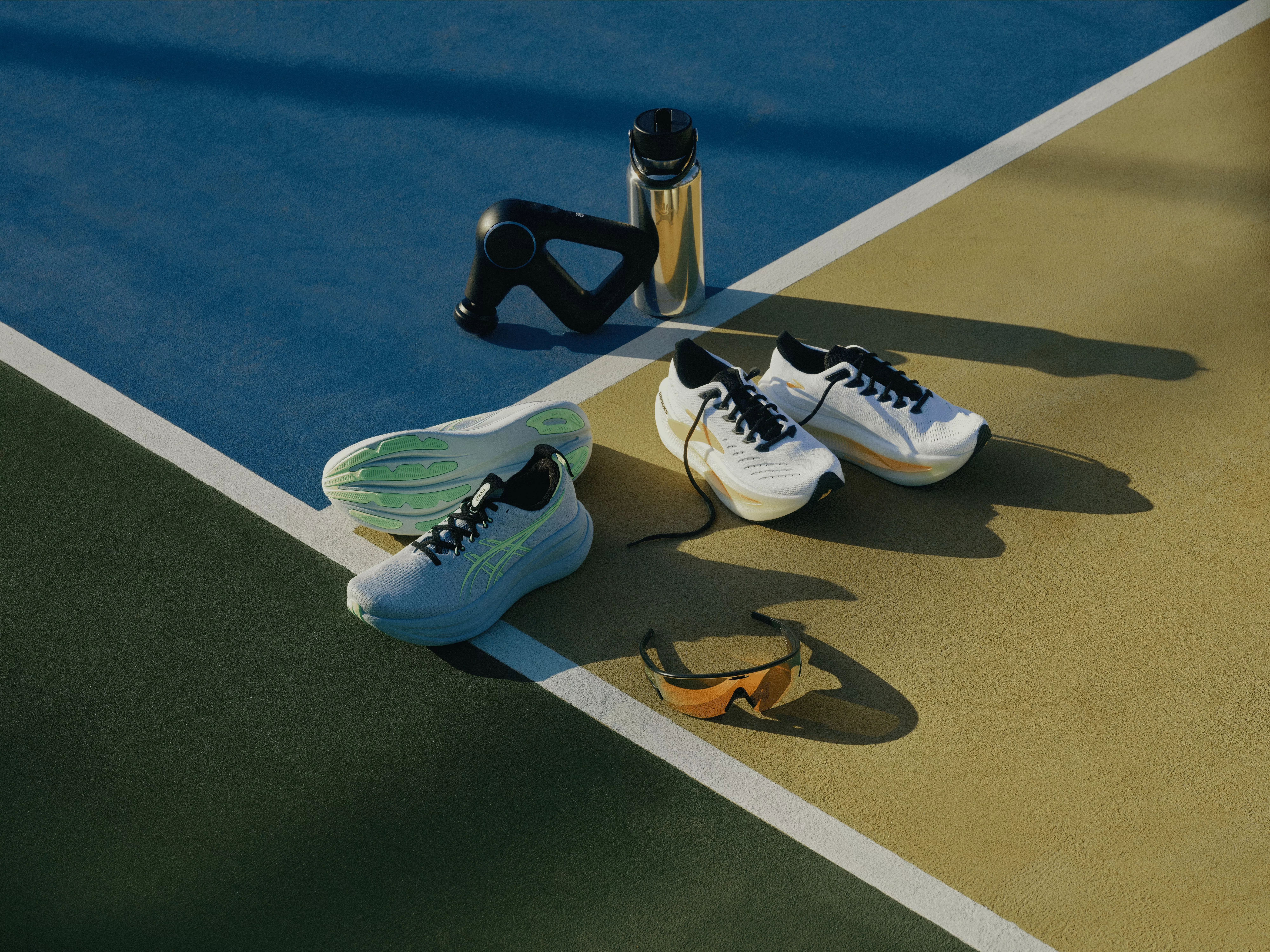 Running shoes, massage gun, water bottle and sports sunglasses laid out on an outdoor tennis court surface