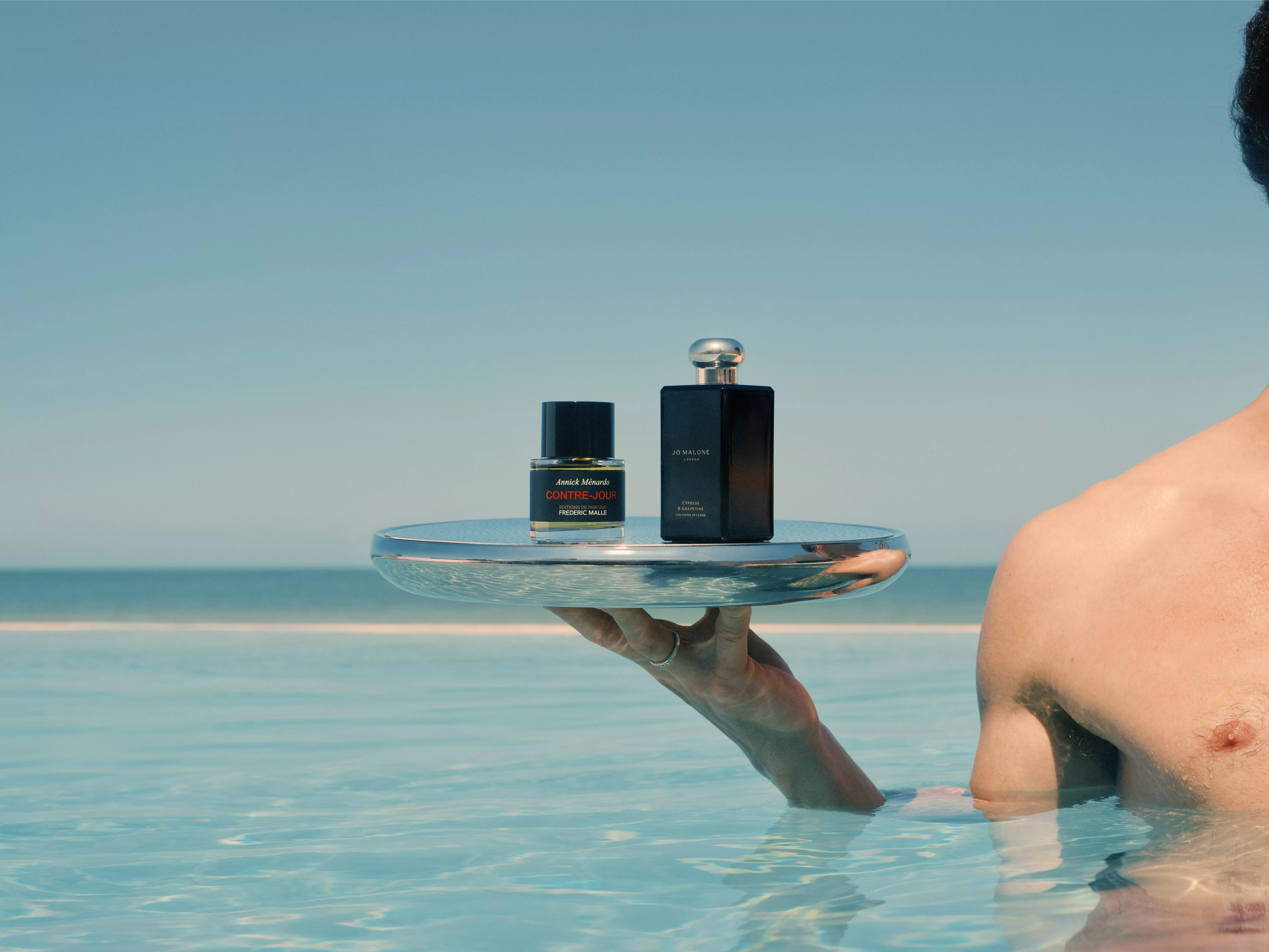Luxury fragrance bottles on a silver tray held above a pool, with sea horizon in the background