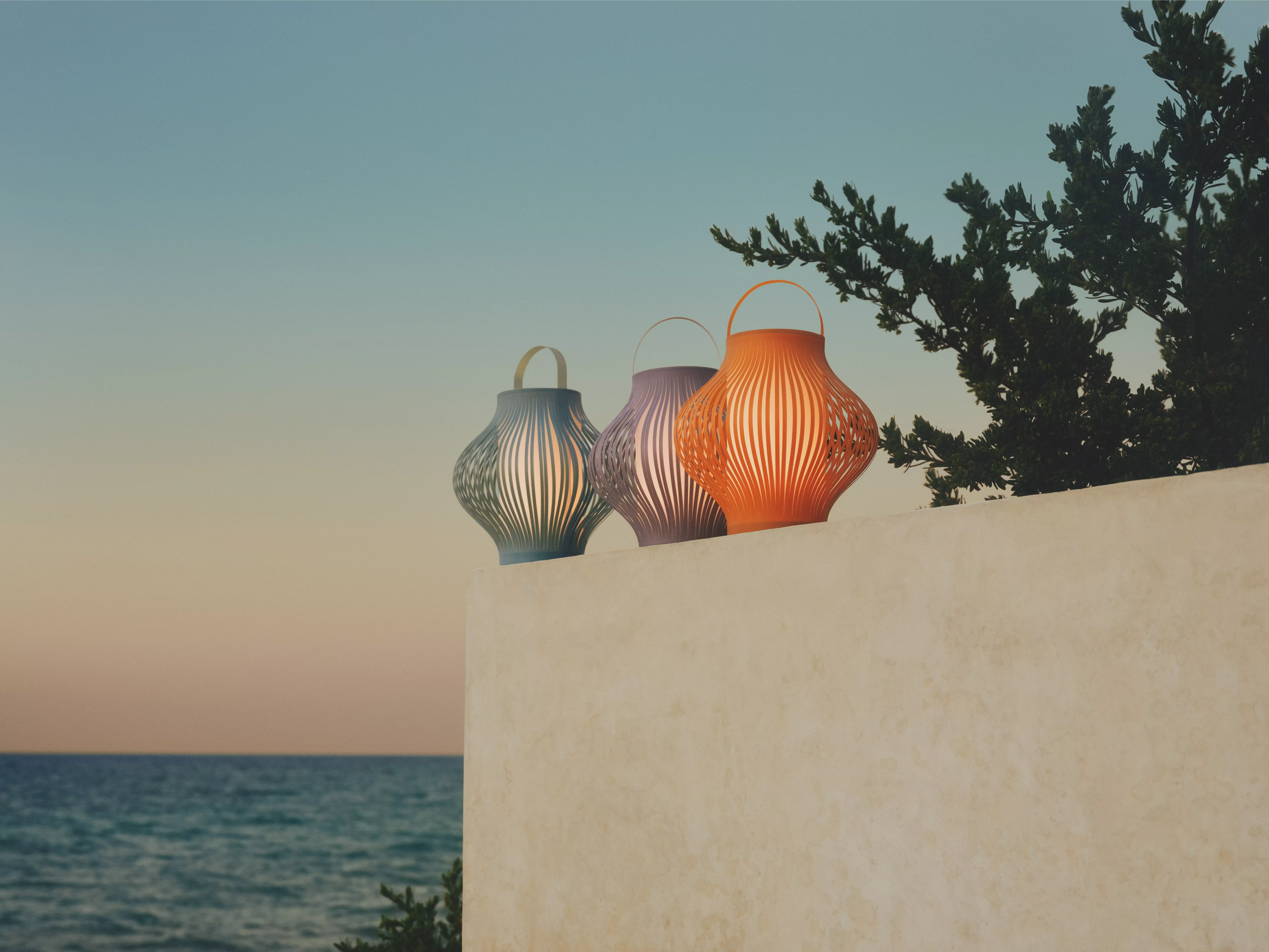 Three colourful outdoor lanterns on a white terrace wall by the sea at sunset, with tree branches overhead
