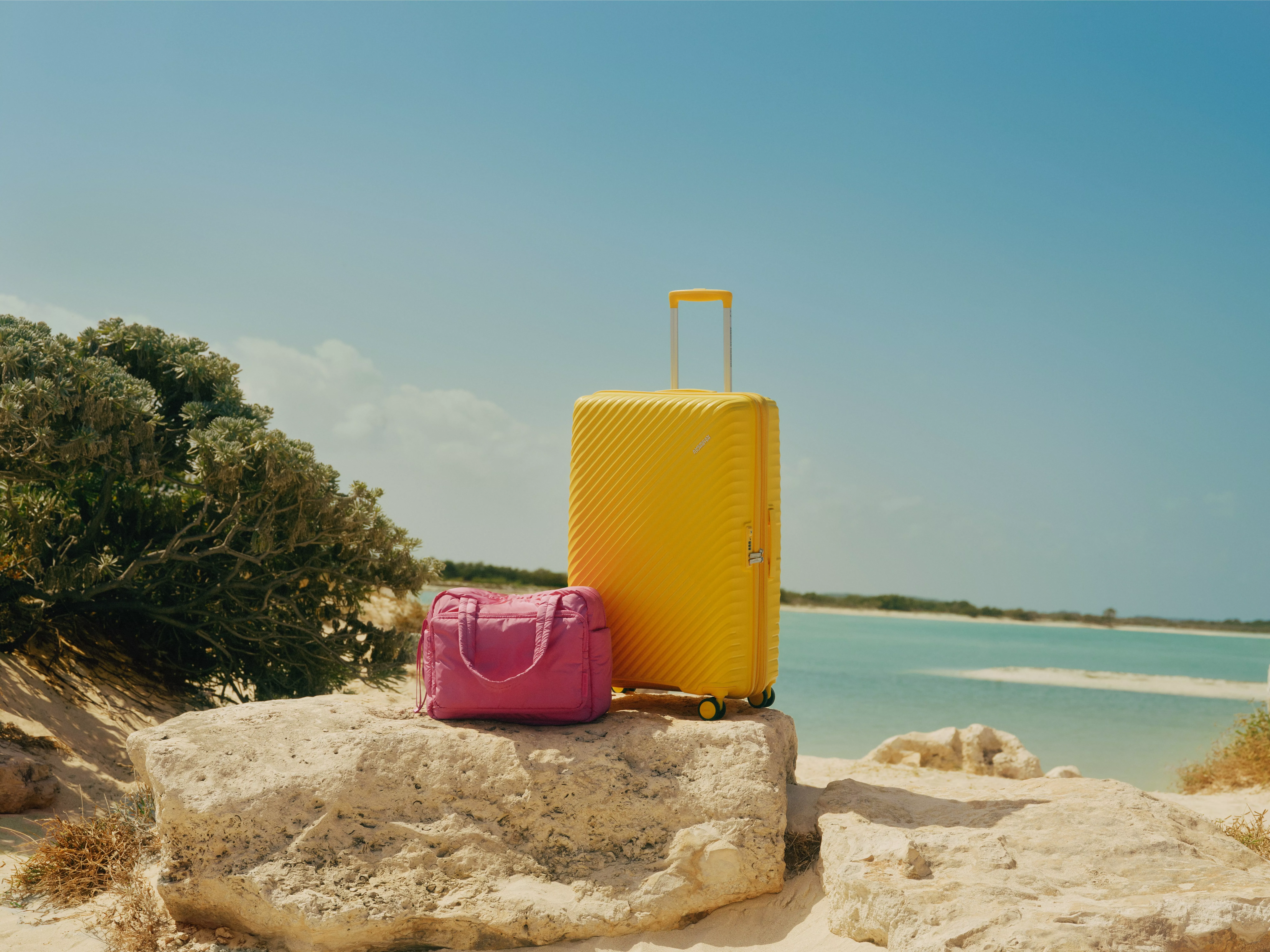 Yellow hard-shell suitcase and pink handbag on rocks by turquoise sea, summer beach travel luggage scene