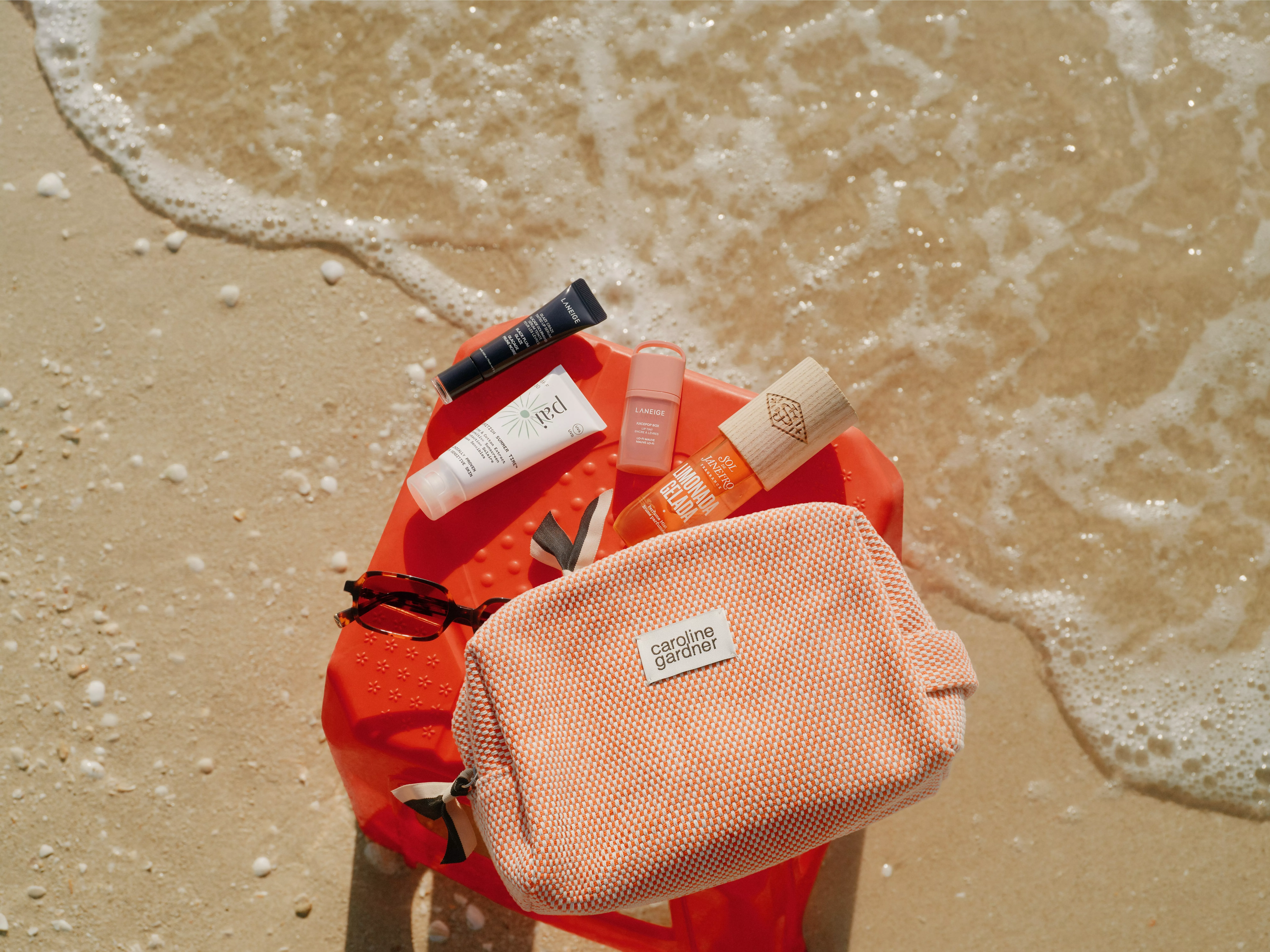 Beach skincare essentials on sand: orange travel pouch, sunglasses, sunscreen and lip balm beside sea foam.
