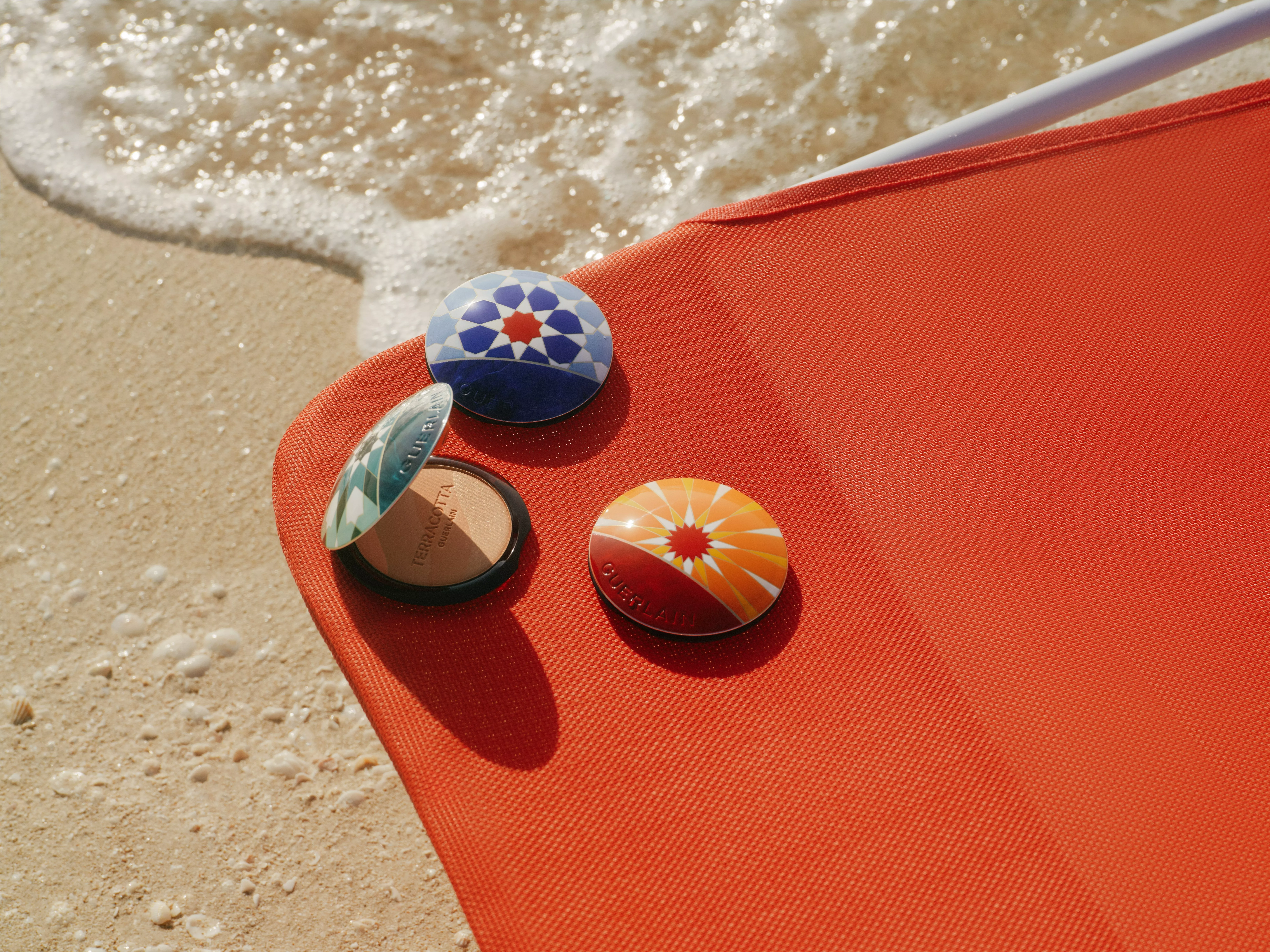 Guerlain bronzing powder compacts on an orange beach lounger beside sea foam on sandy shoreline in sunlight