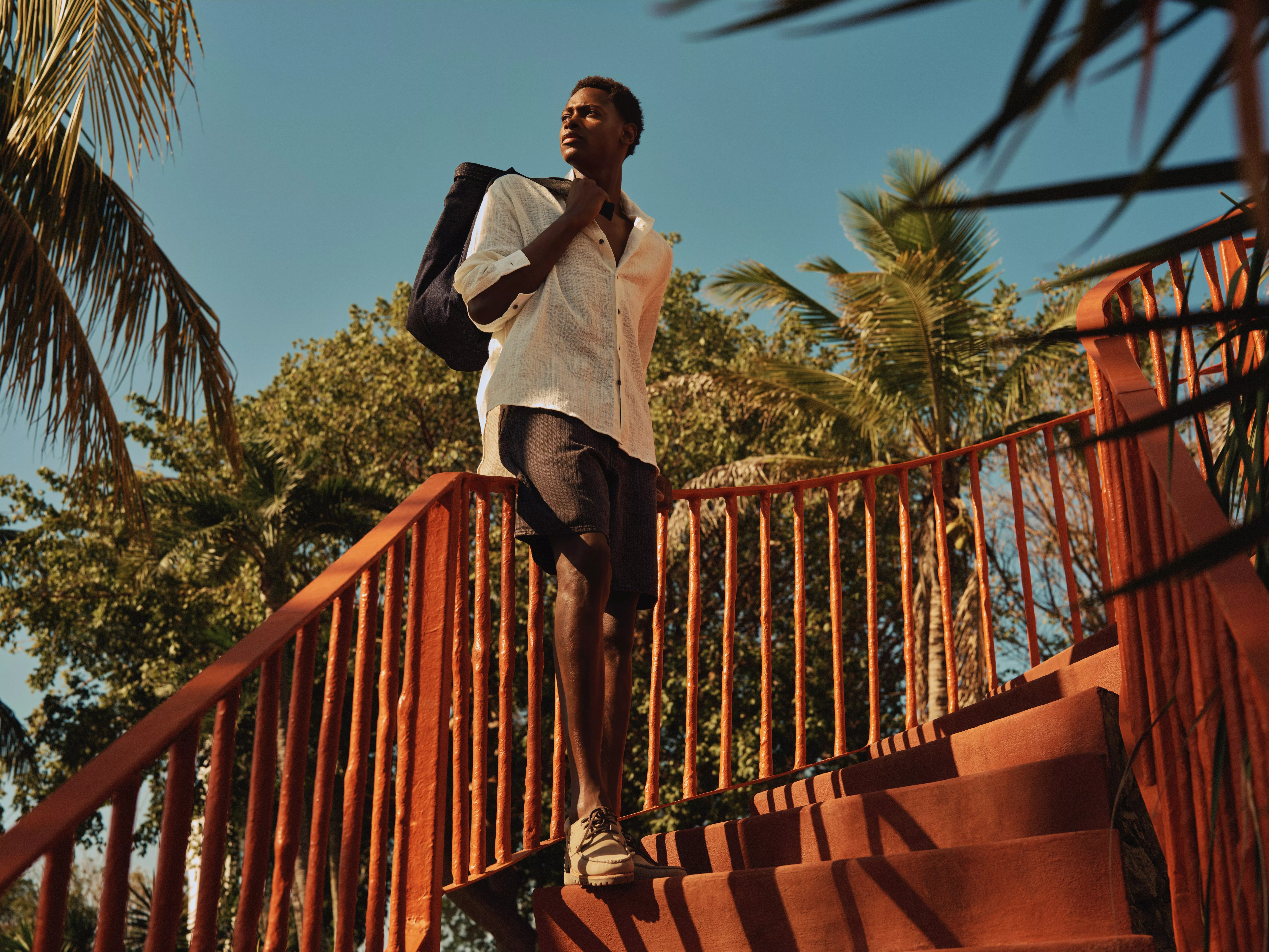 Young man with backpack walking down red outdoor steps, framed by palm trees under clear blue sky