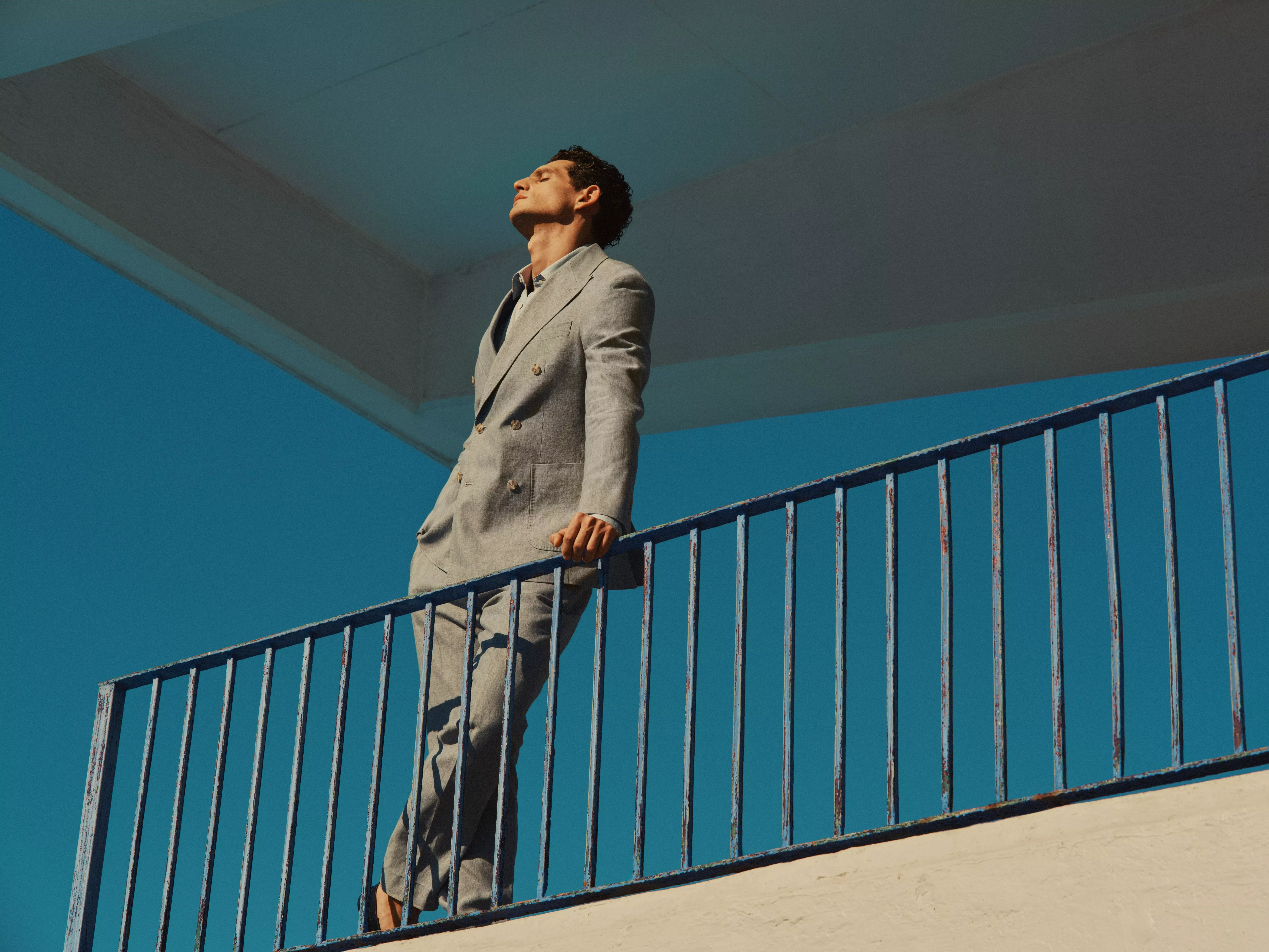 Man in light grey suit standing on outdoor stair balcony, leaning on blue rail under modern architecture, sunny sky