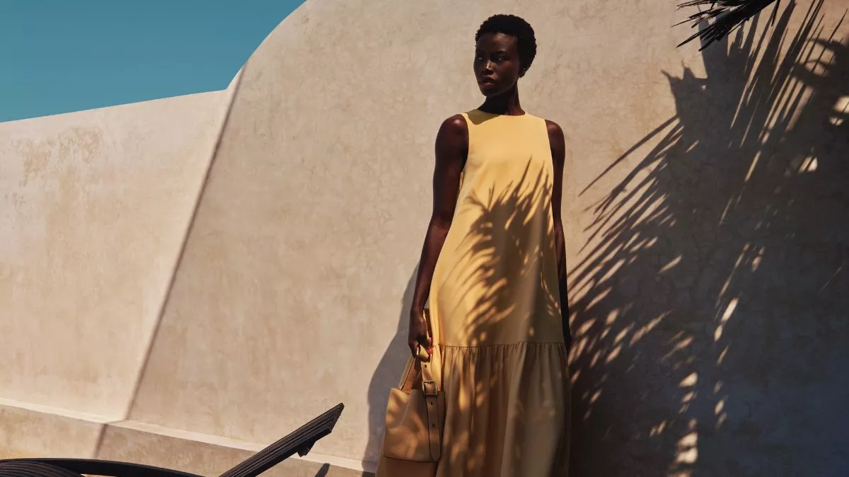 Fashion model in a pale yellow maxi dress holding a handbag against sunlit stucco wall and palm shadows
