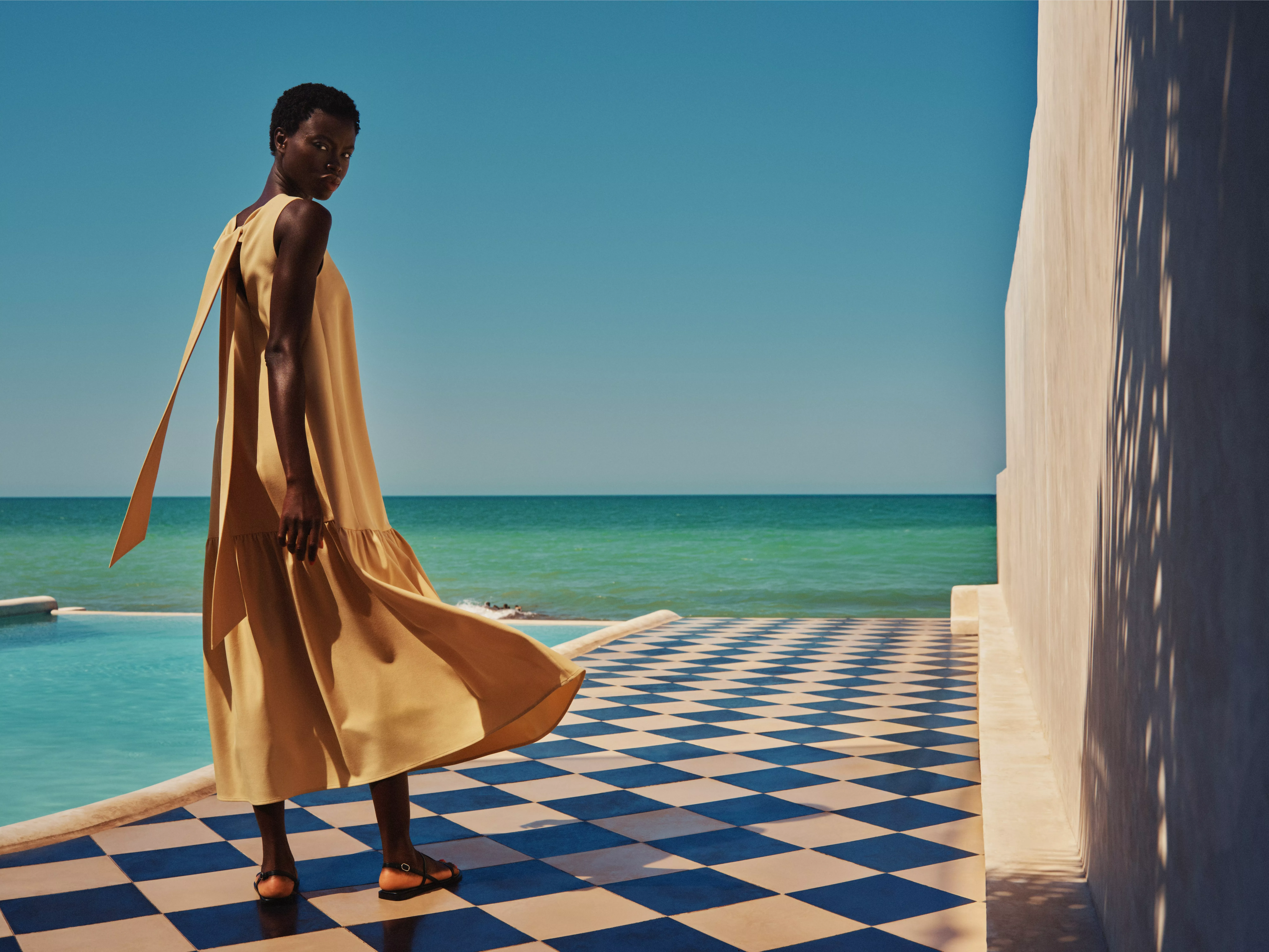 Fashion model in flowing mustard maxi dress by poolside, blue checkered terrace, seaside backdrop in sunlight