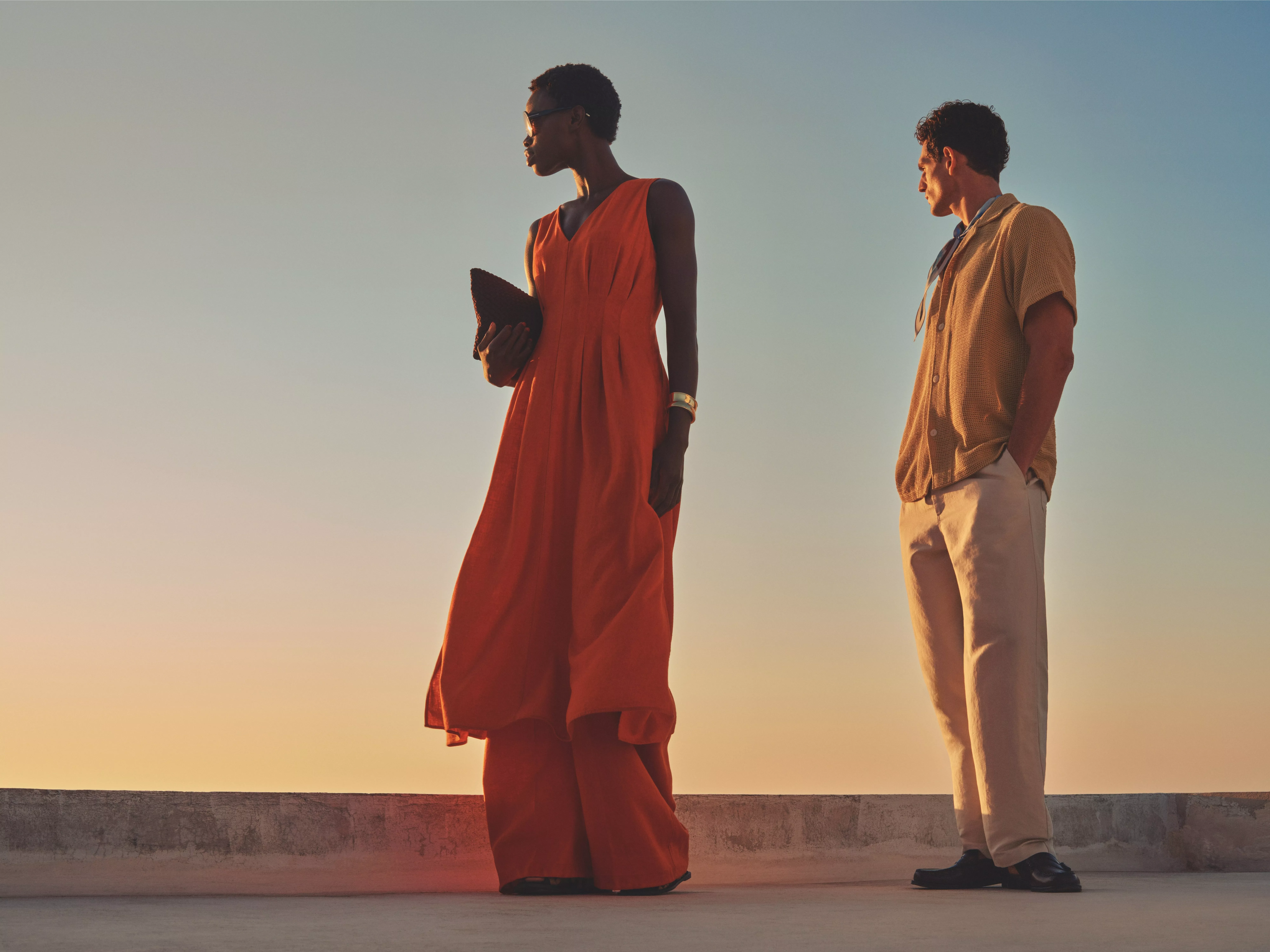 Stylish couple in summer outfits at sunset, woman in orange maxi dress holding clutch, man in beige trousers