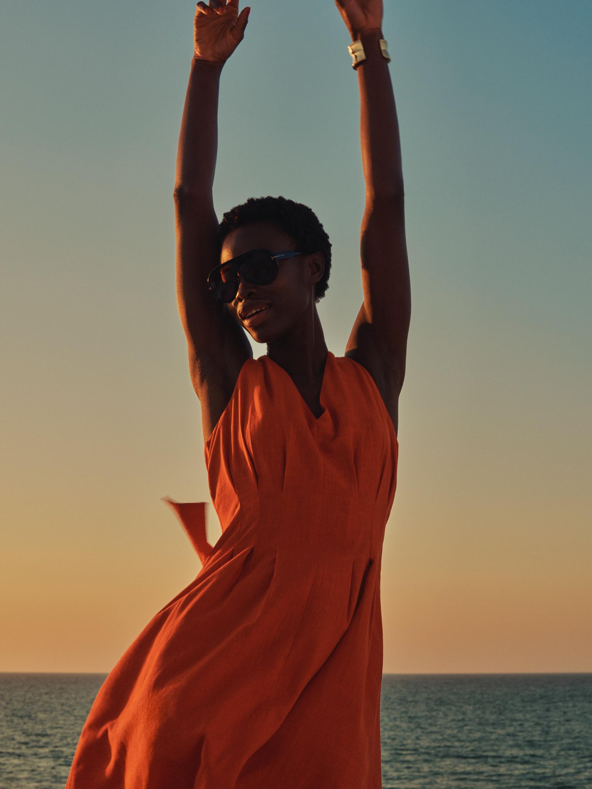Woman in orange summer dress and sunglasses stretching arms by the sea at golden hour sunset sky
