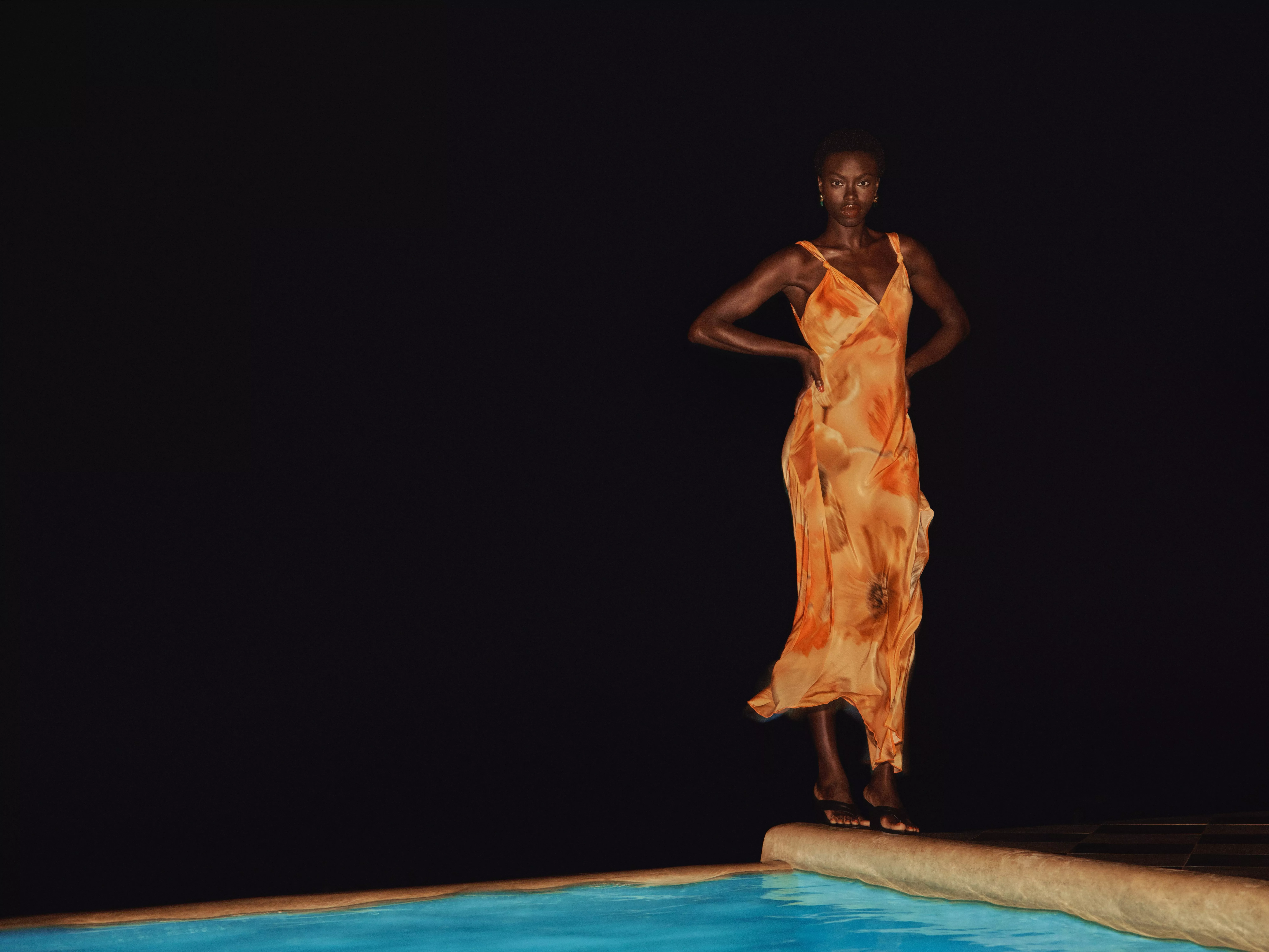 Fashion model in Floral orange satin slip dress standing poolside at night against dark background, editorial style