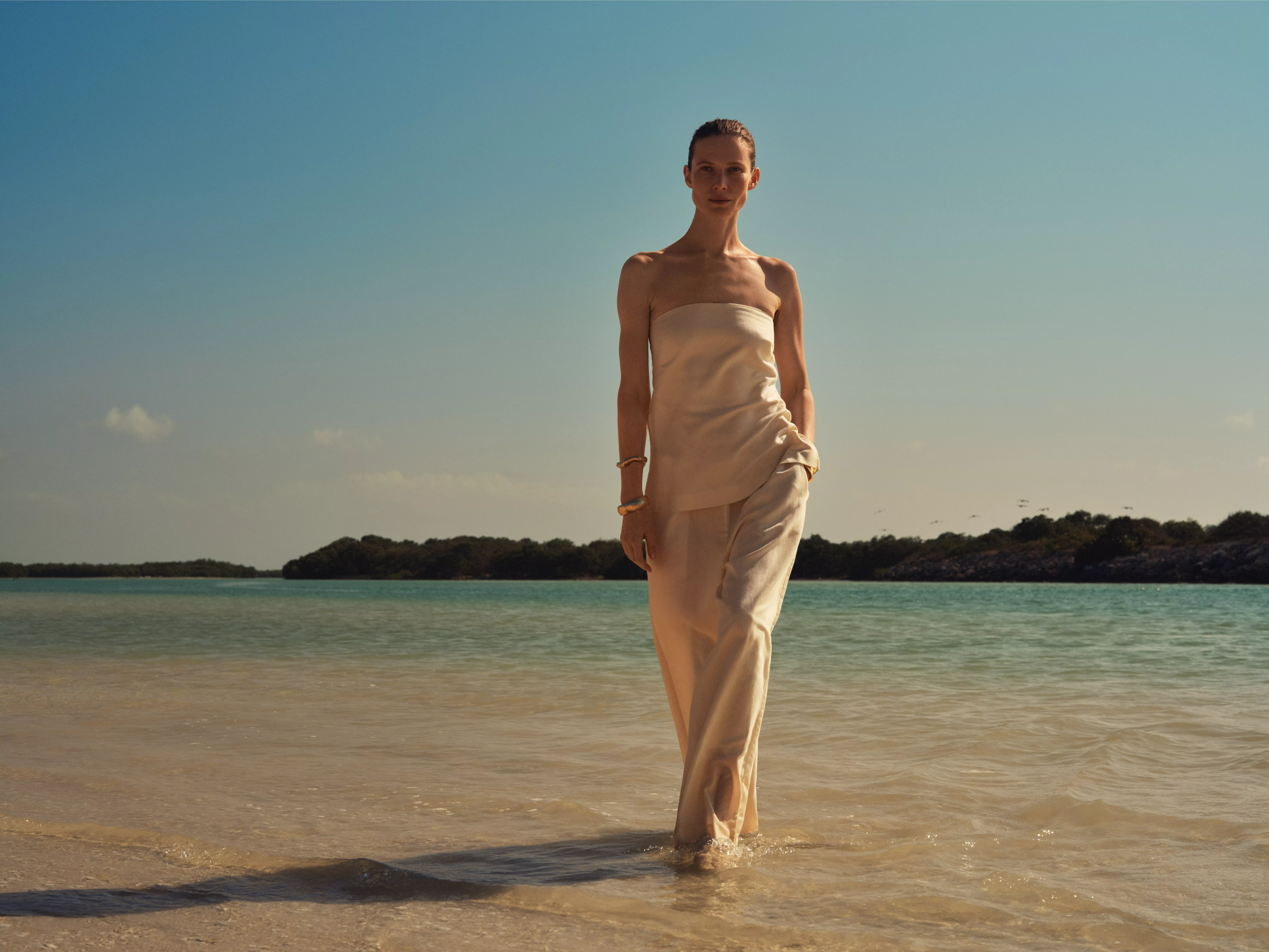Woman in elegant cream dress walking in shallow sea on tropical beach, blue sky and island horizon backdrop